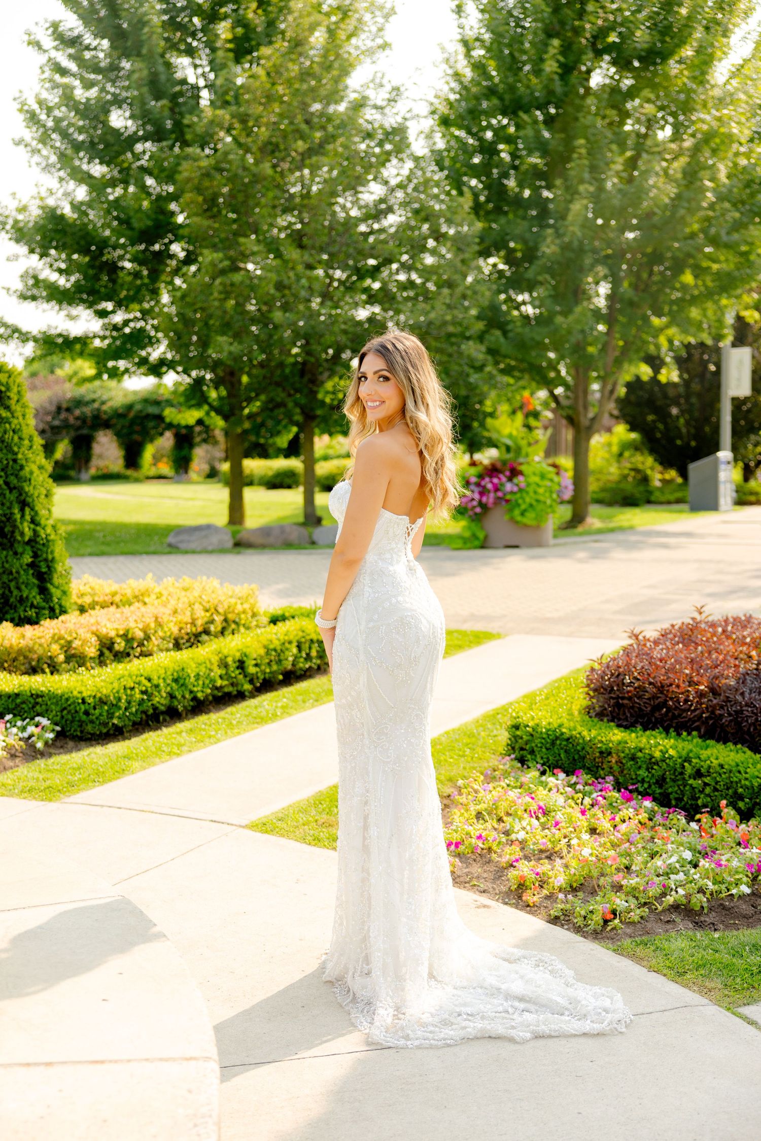 Bride in fitted lace wedding gown poses in garden setting with manicured landscaping.