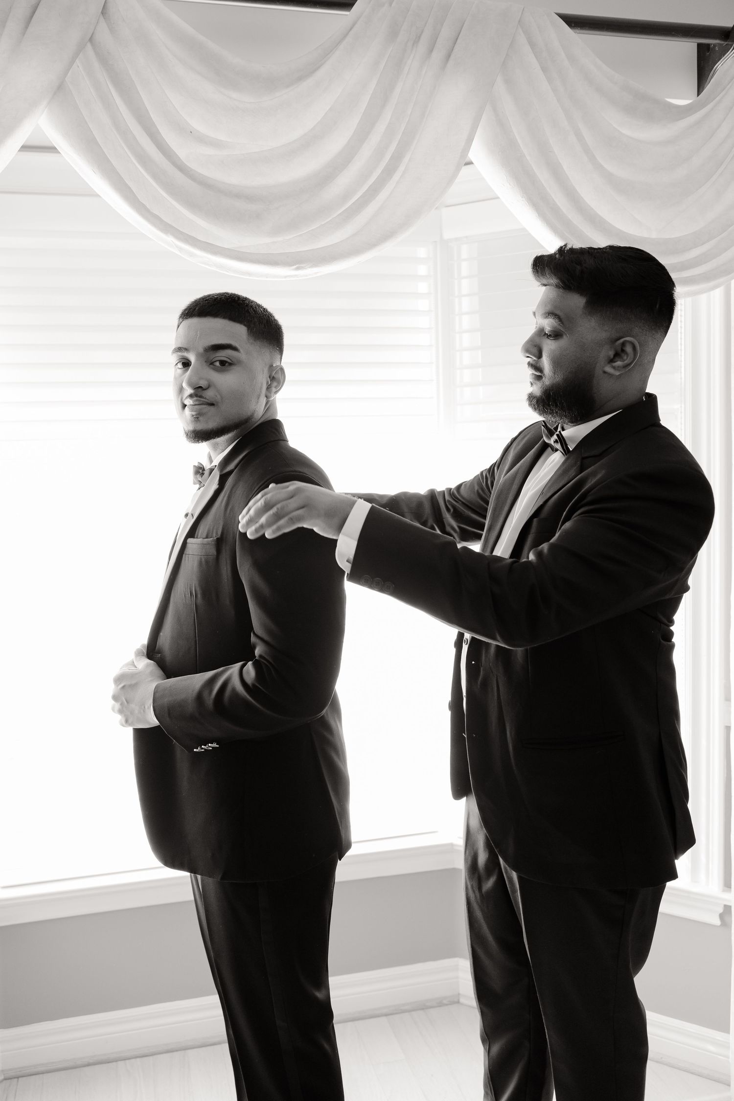 Two people in formal wear make final adjustments to clothing in a room with draped curtains.