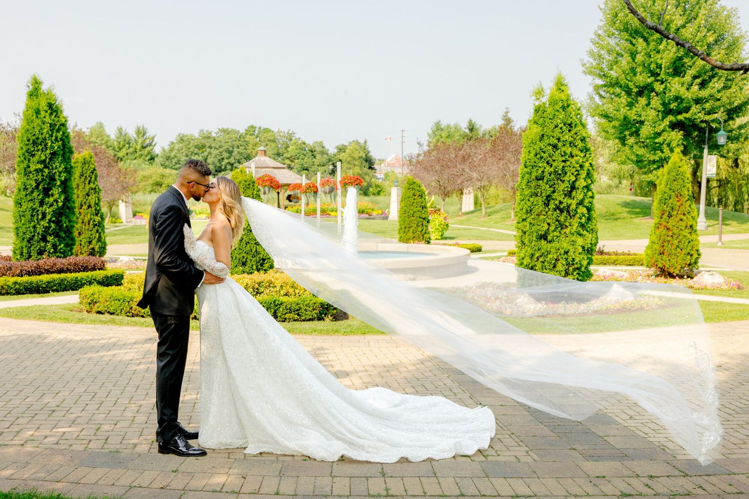 Elegant wedding photo showcases couple on brick path with dramatic flowing veil and garden backdrop.