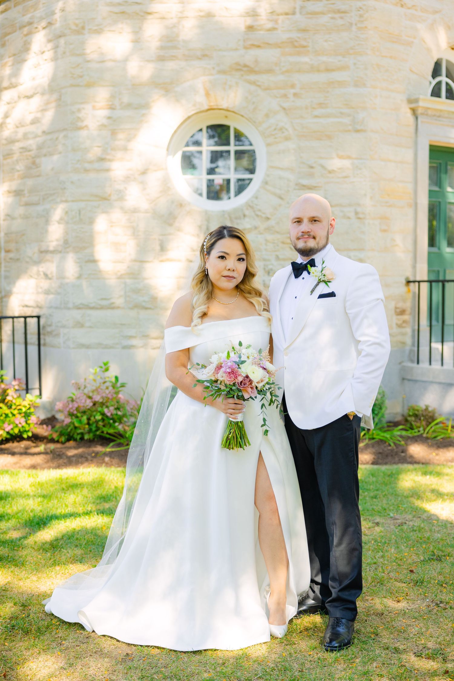 A wedding couple poses in front of a stone building with a circular window and green door.