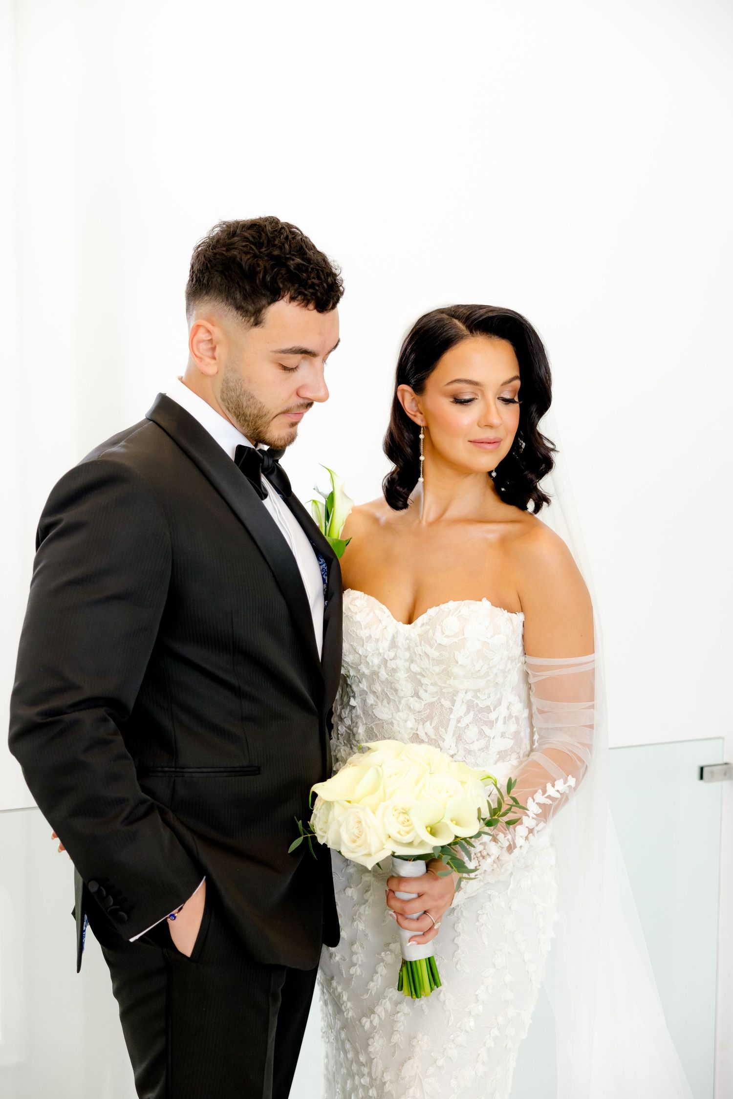 Elegant wedding couple posing with white roses against bright white backdrop.