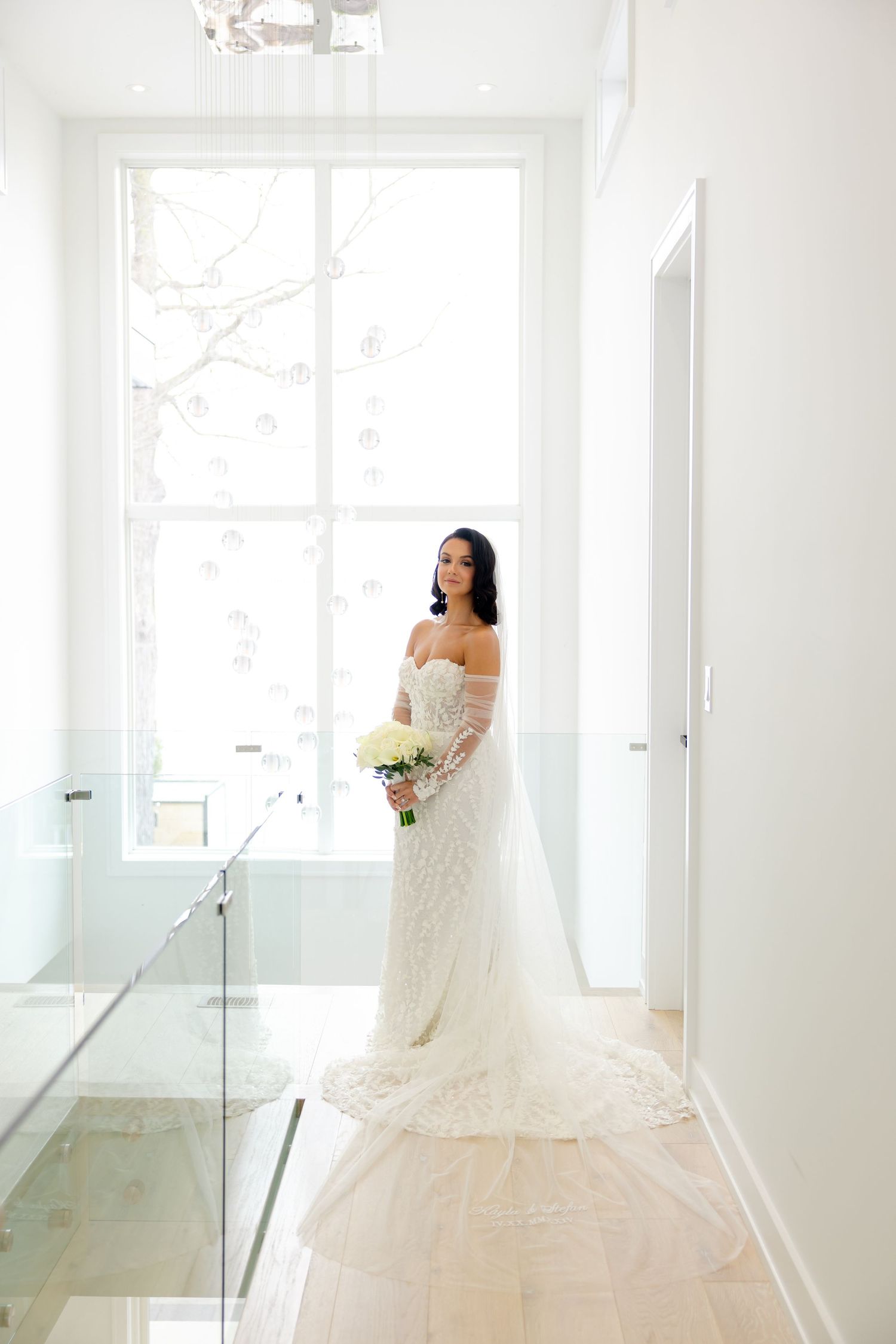 A romantic bridal portrait by a large window showcasing an off-shoulder lace gown with flowing train.
