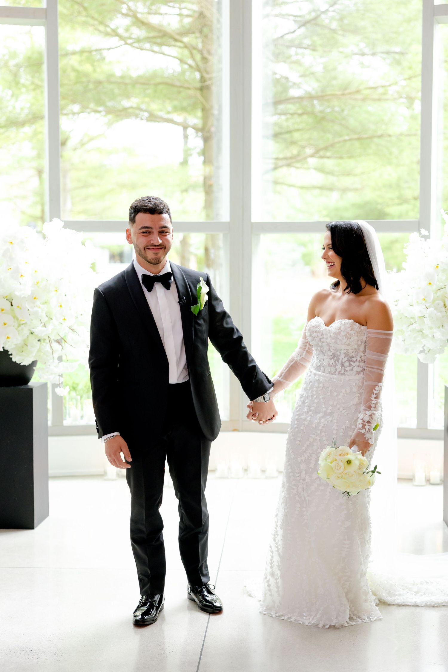 A couple in formal wedding attire holding hands in an elegant room with white flowers and natural lighting.