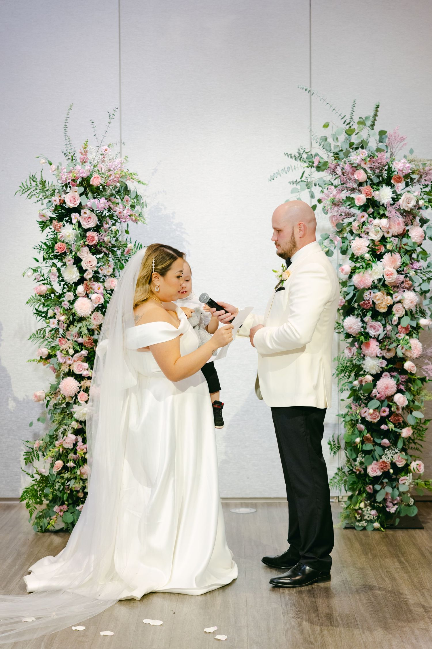 Couple exchanges rings between pink floral arrangements during wedding ceremony.