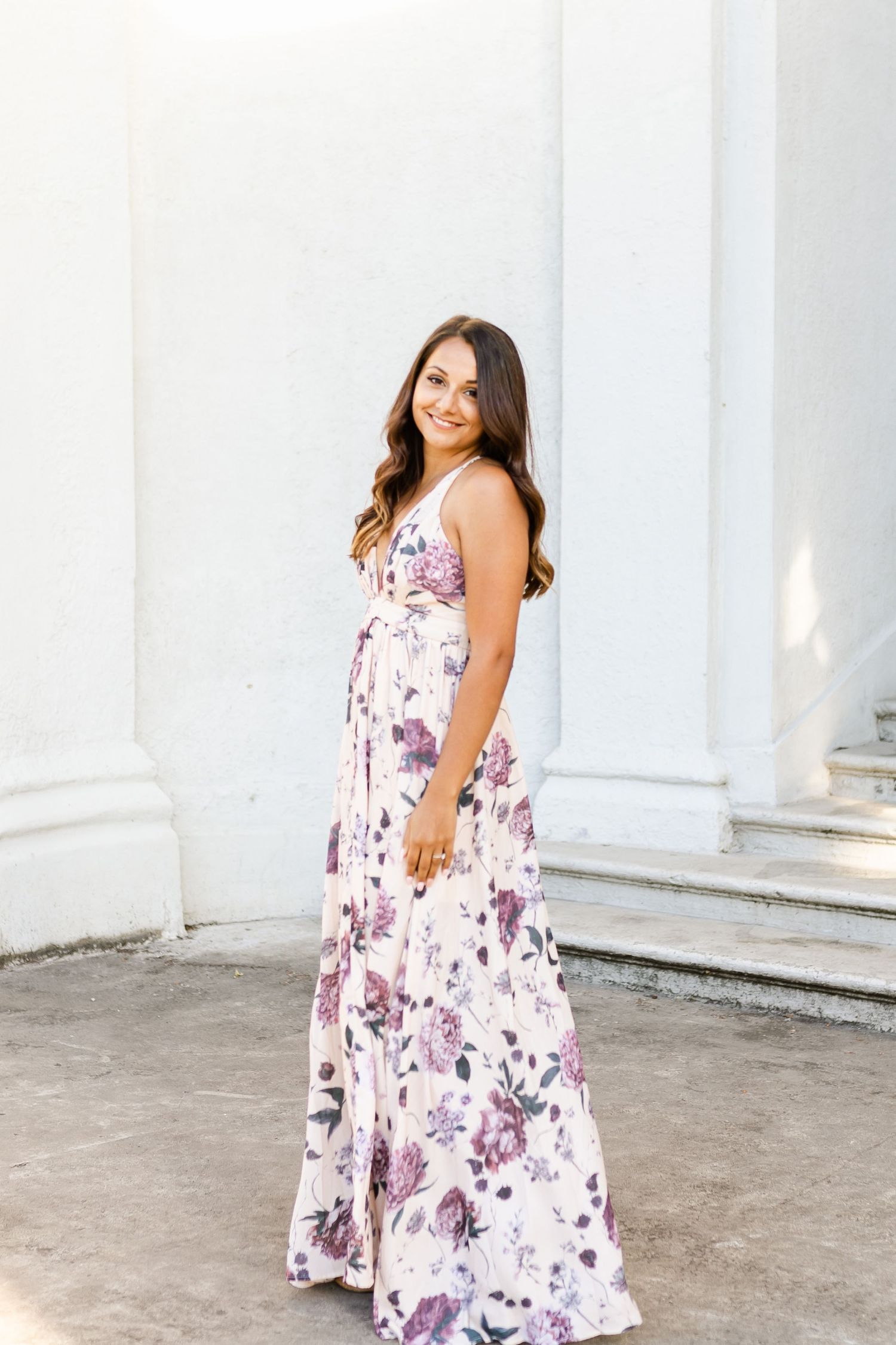 Woman in sleeveless floral maxi dress smiling against white architectural backdrop.