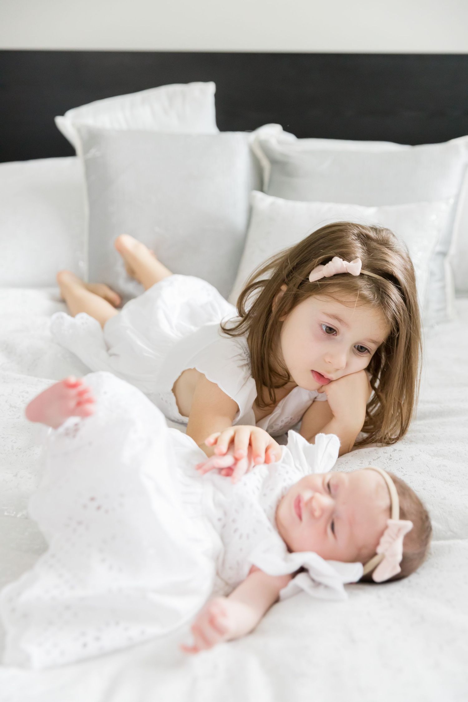 Two children in white clothing lying on white bed looking at each other.