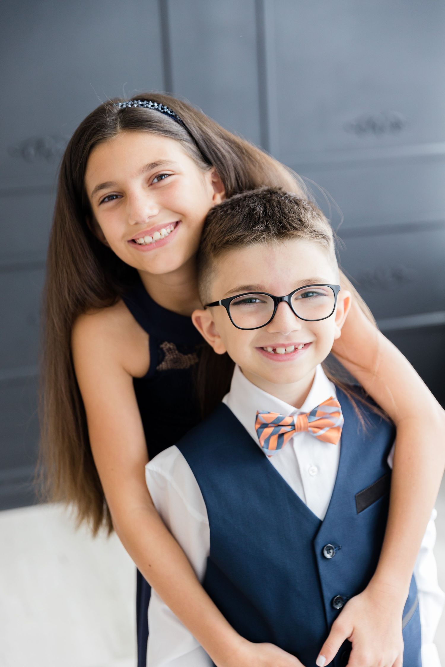 Sweet portrait of siblings embracing in formal navy outfits against a classic gray backdrop.
