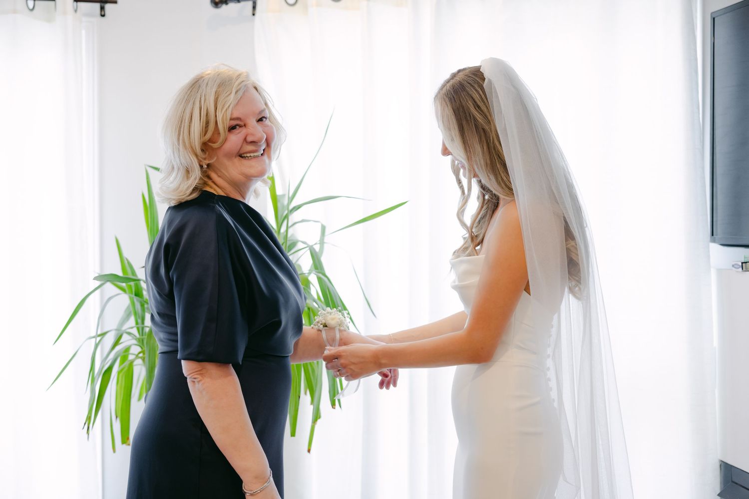 Joyful interaction between bride in white gown and woman in navy dress by window.