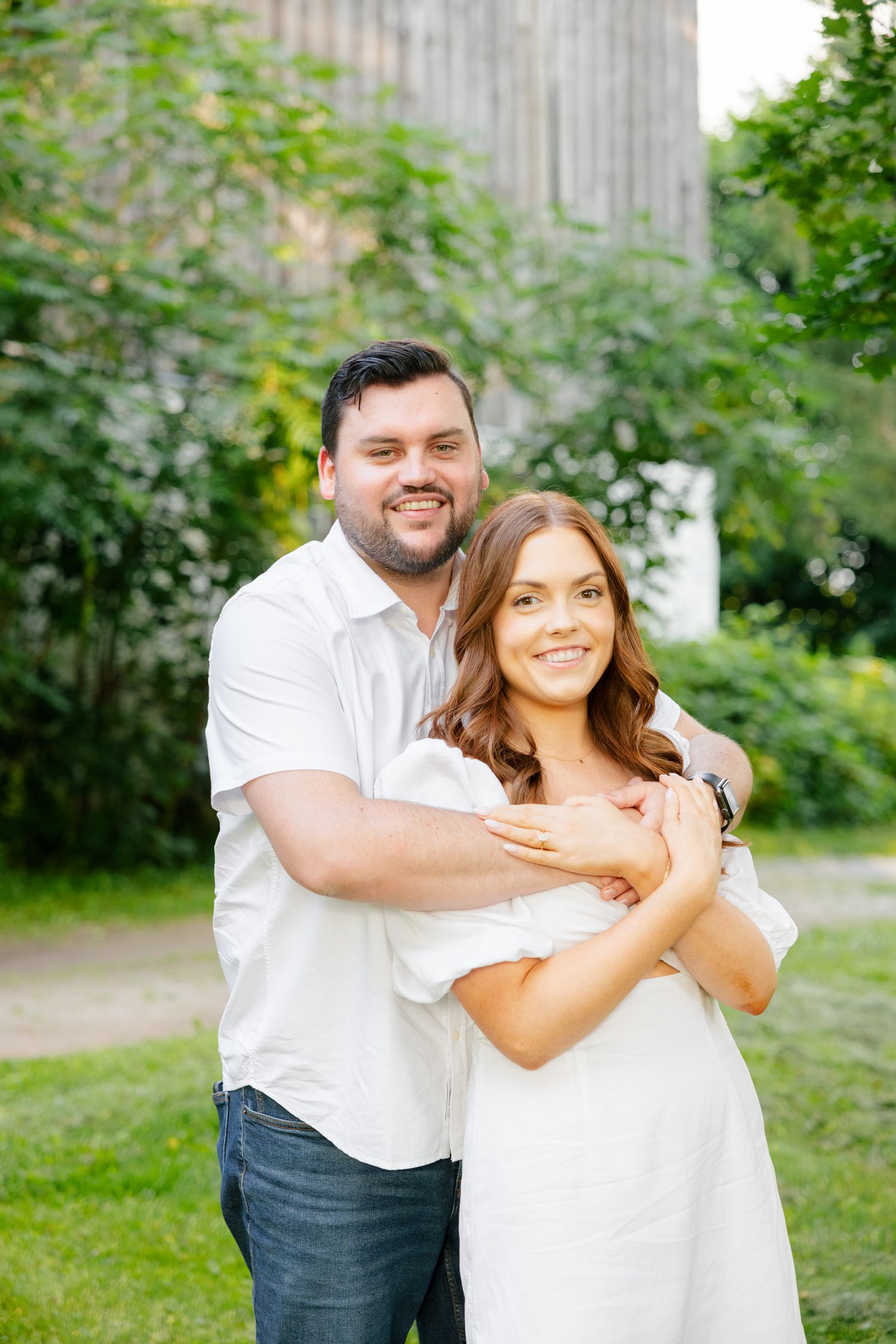 A couple shares an intimate embrace while standing on grass near flowering bushes.