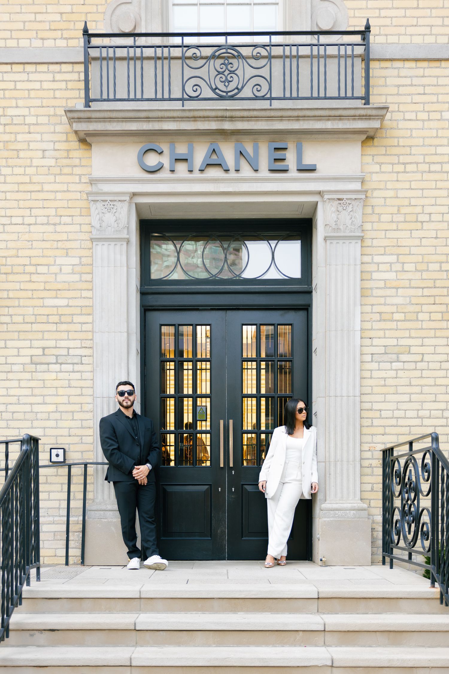 A couple in coordinated outfits stands before the elegant entrance of a Chanel boutique.