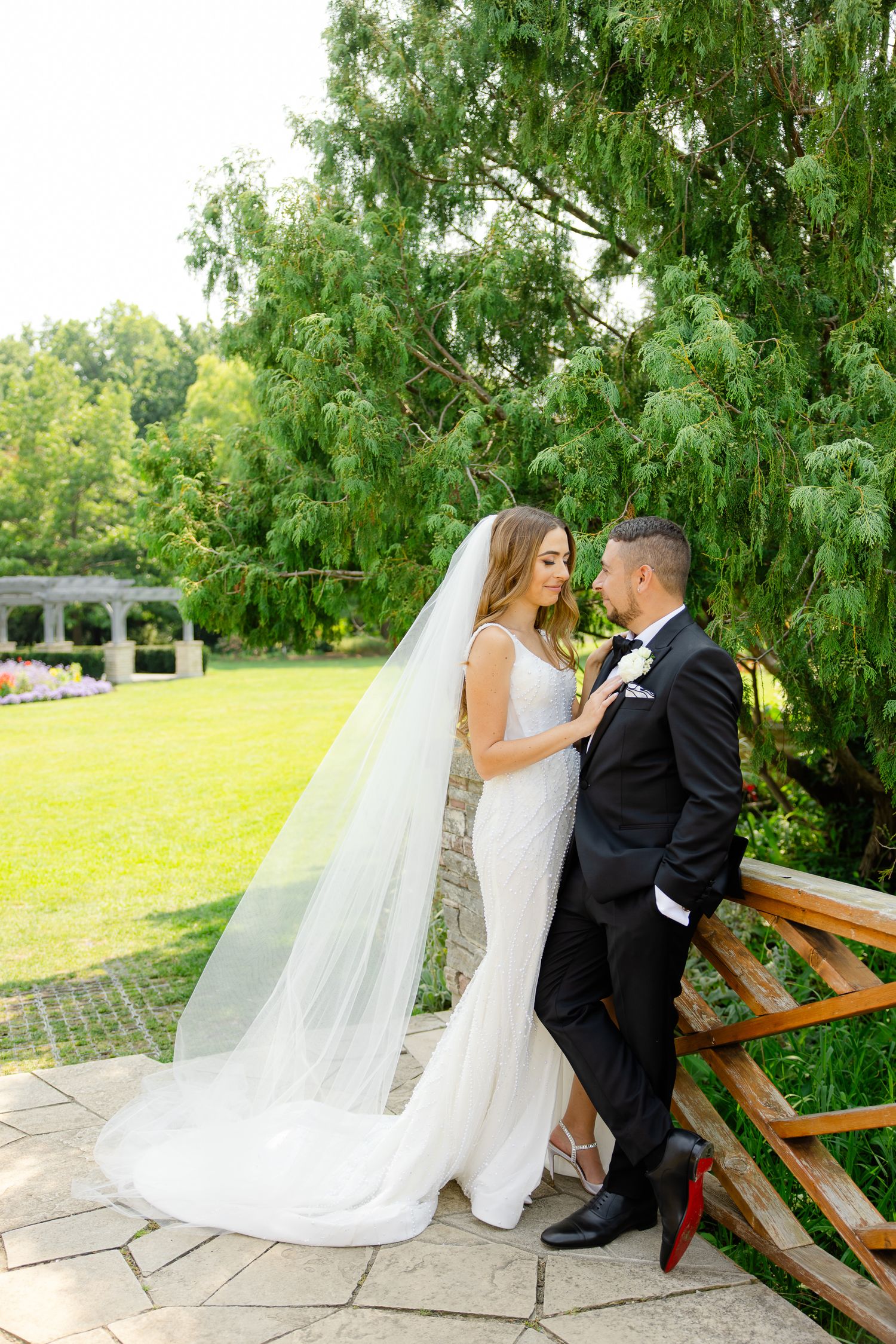 Romantic garden wedding photo with couple standing by wooden railing under weeping willow tree.
