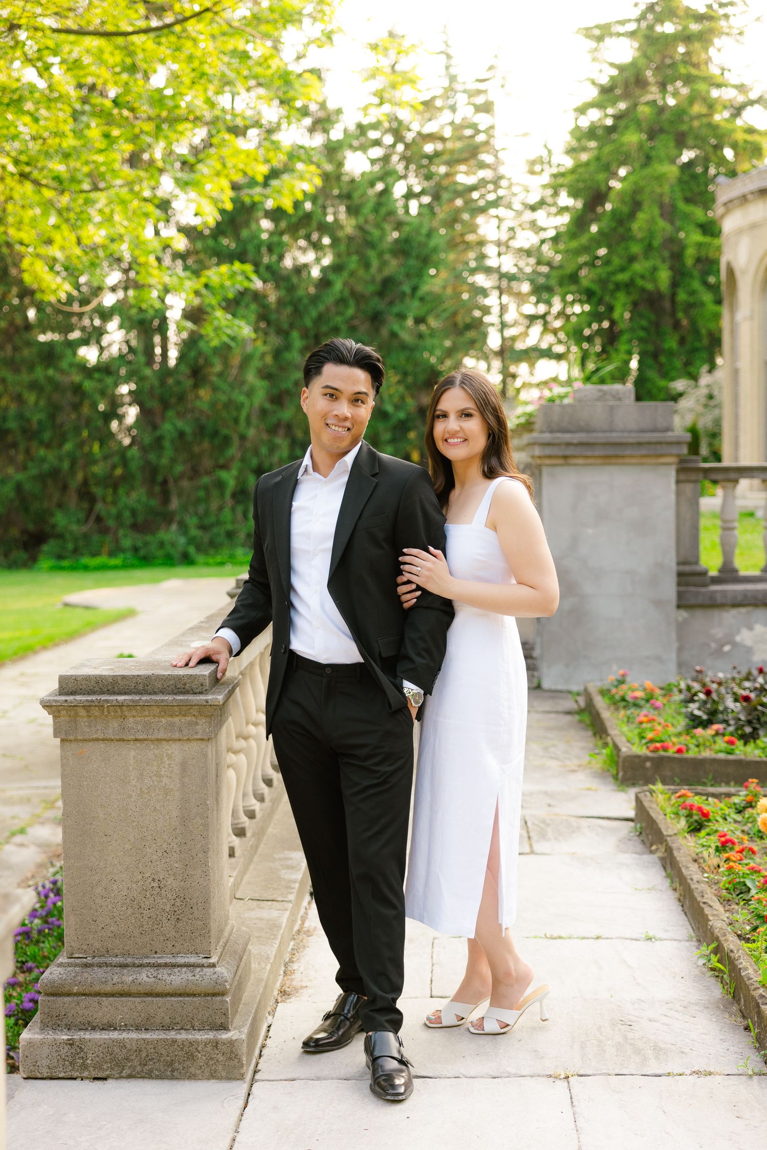 Elegant couple poses by stone railing in formal garden with blooming flowers.