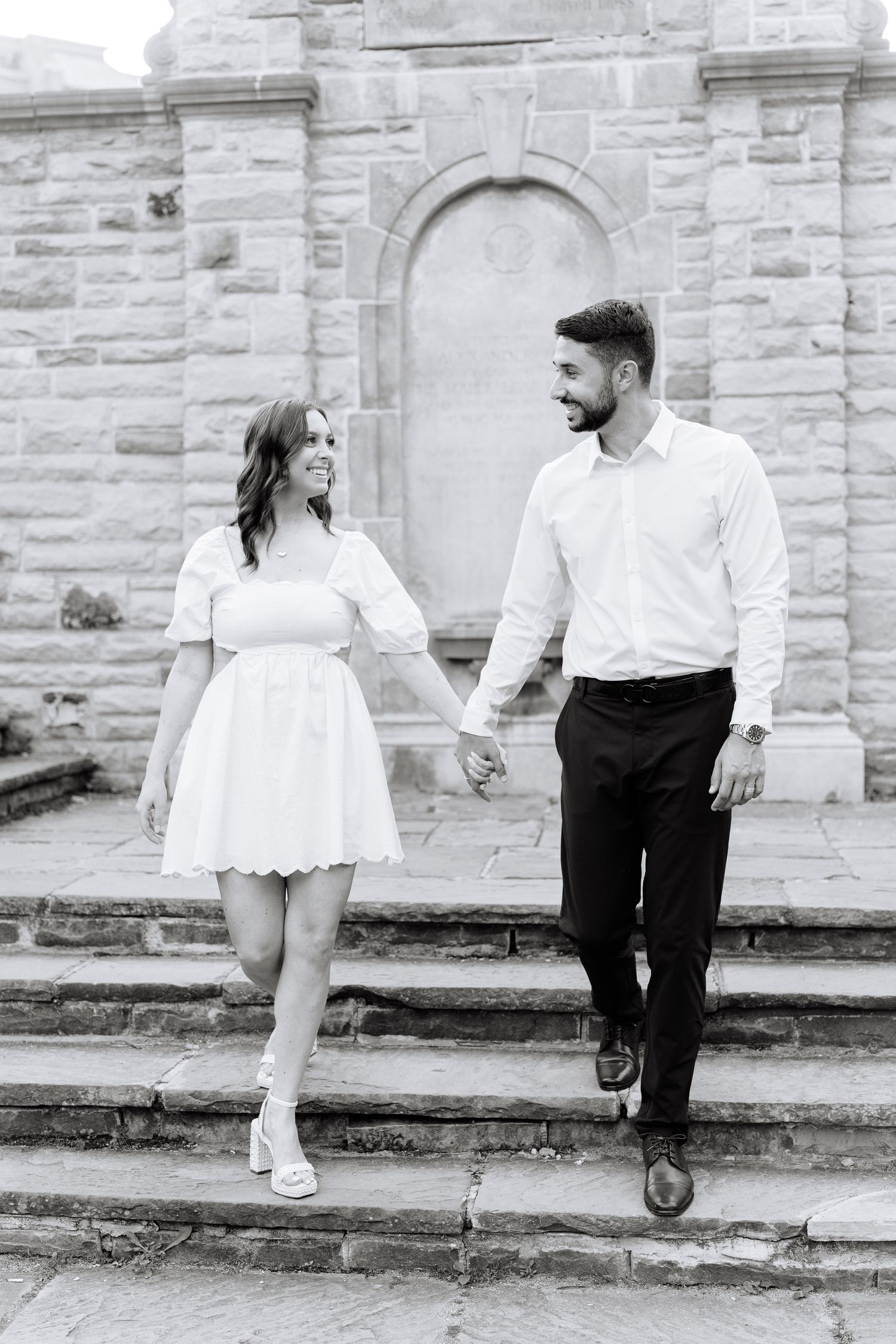 A couple descends stone steps in matching white outfits at a historic stone building.