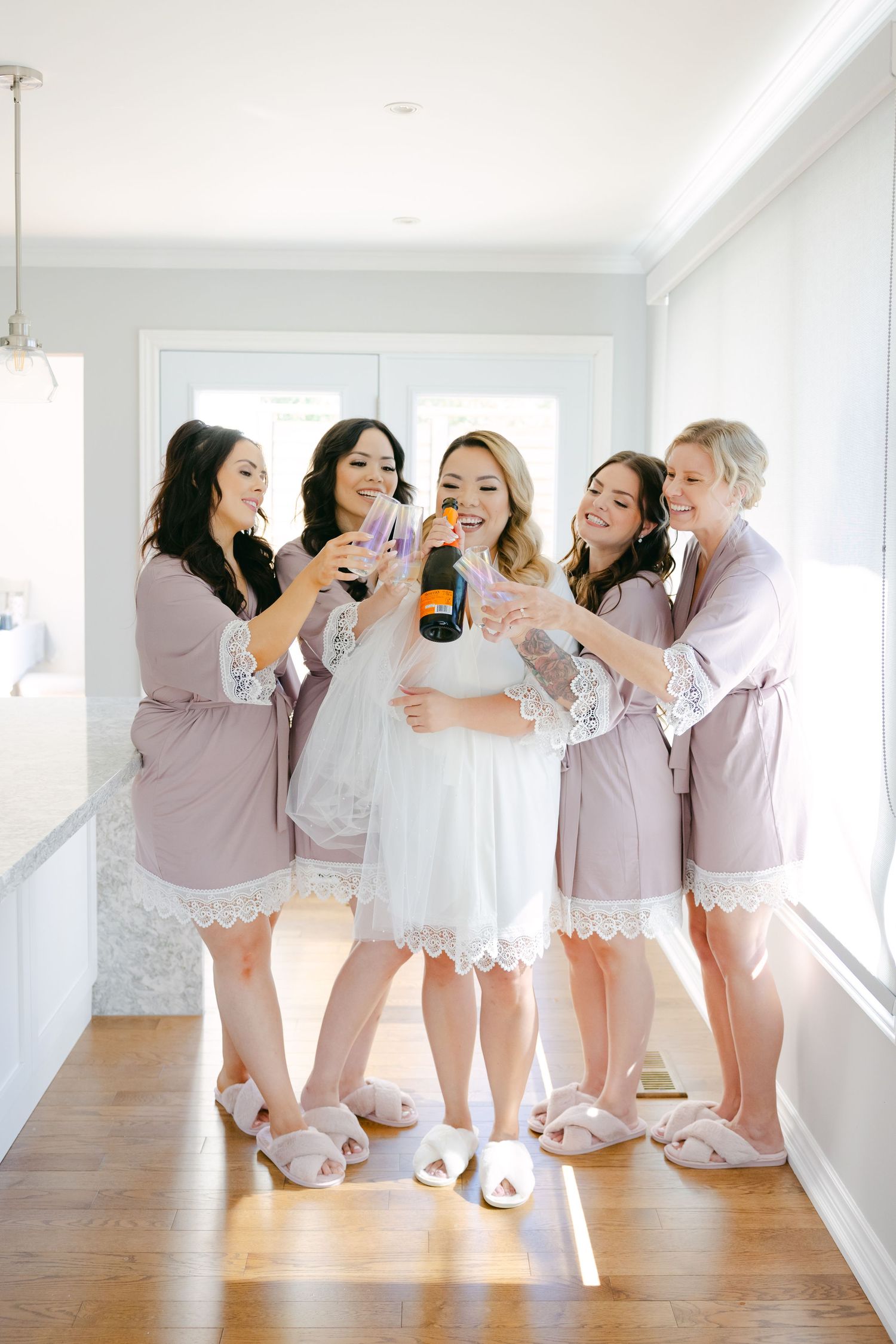 Bridal party in matching mauve robes sharing champagne and laughter.