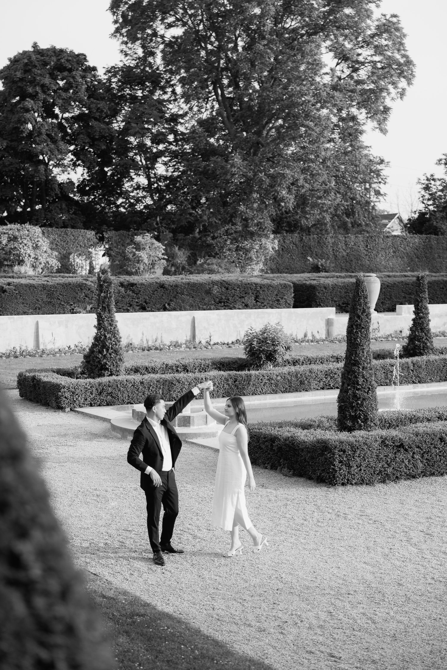 A black and white photo shows two people dancing in a formal garden with cypress trees and hedges.