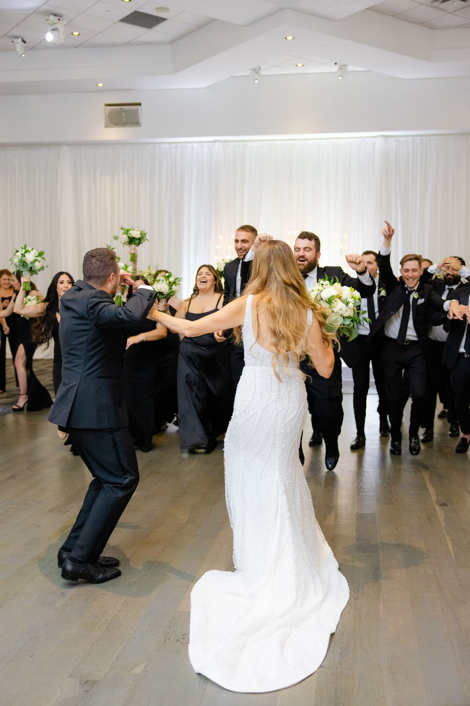 Wedding party celebrates with dynamic dance moves in modern reception venue with white drapes.
