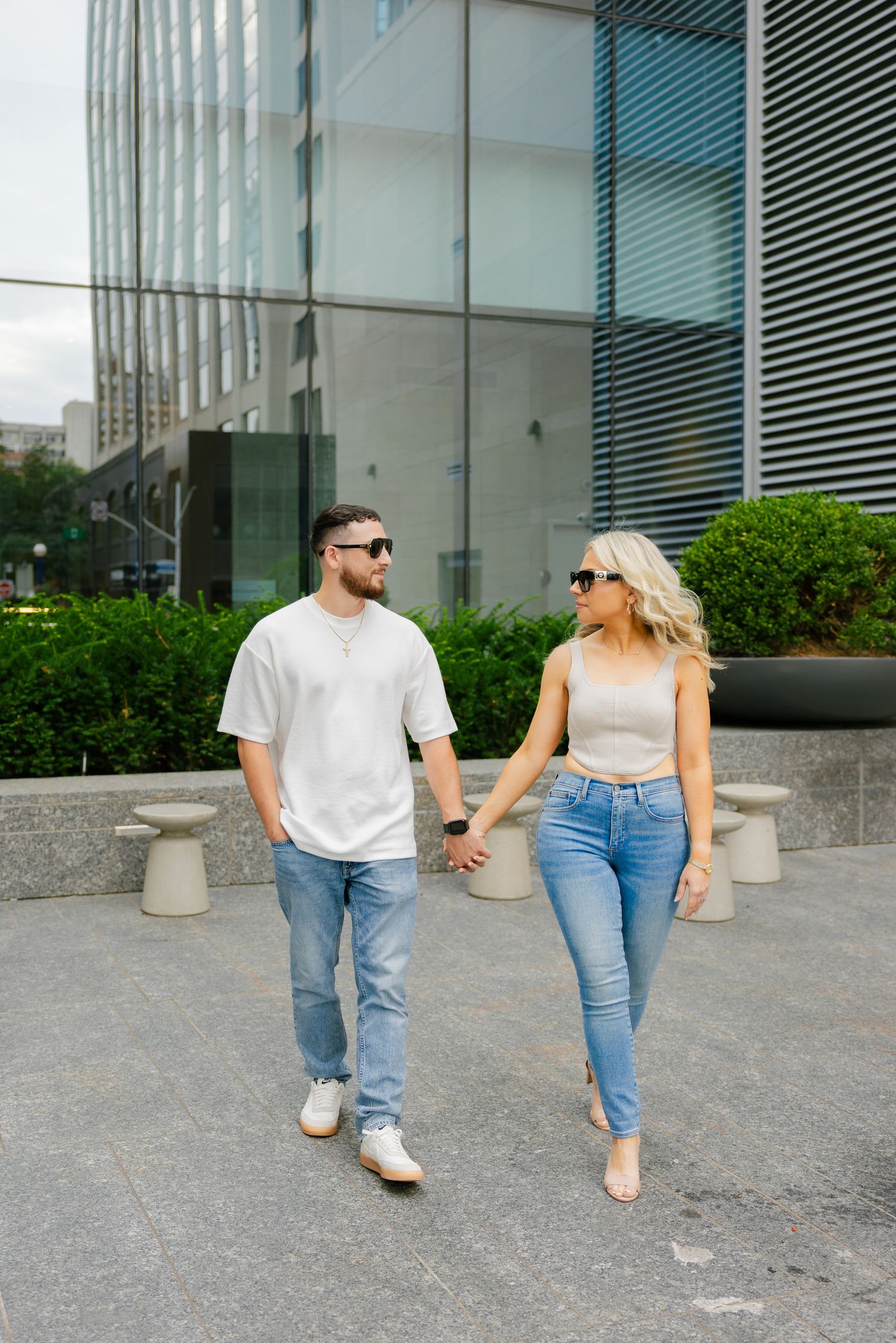 A couple in casual summer attire stroll together near landscaped bushes.