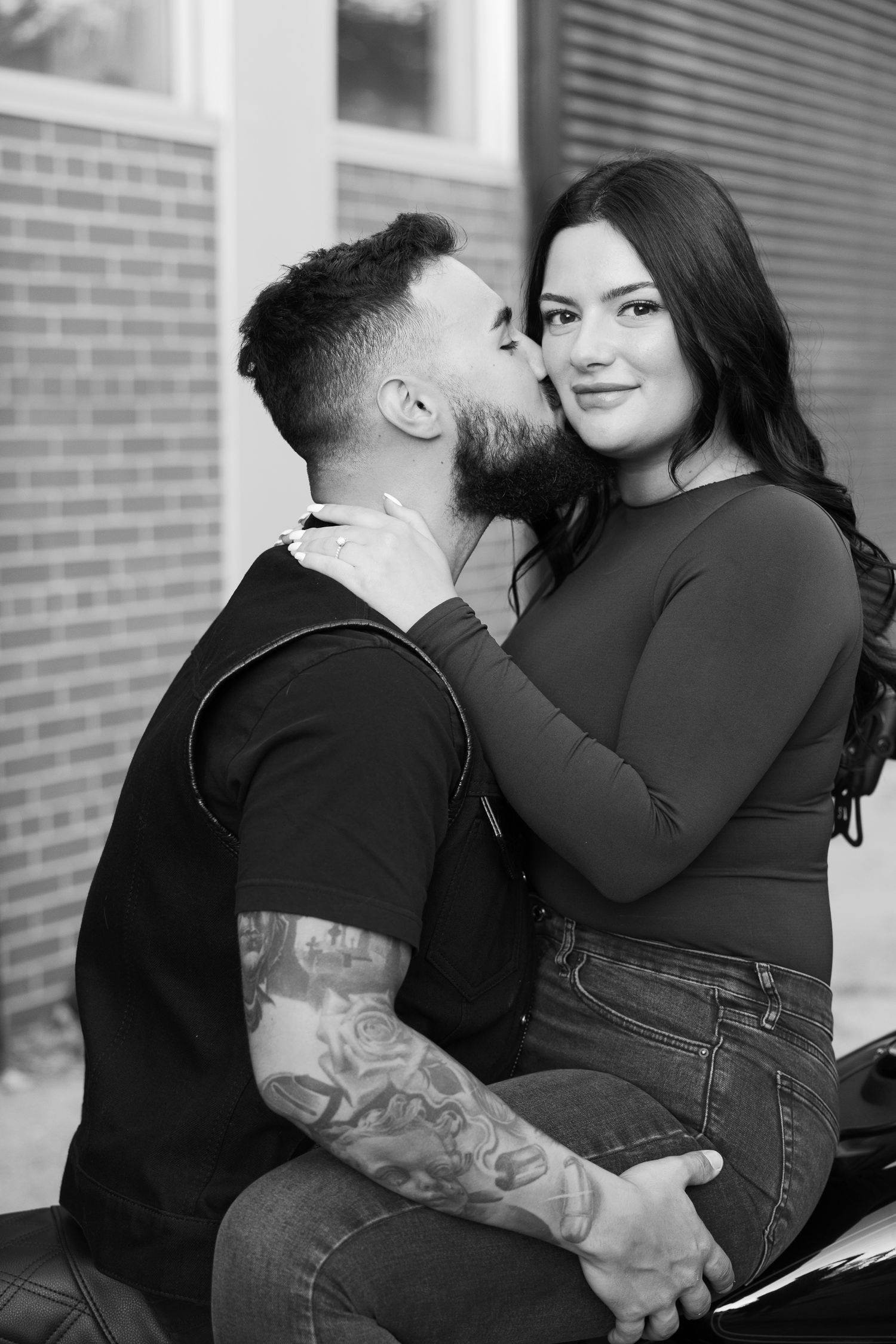 A romantic couple shares an intimate moment while sitting against a brick wall in black and white photography.