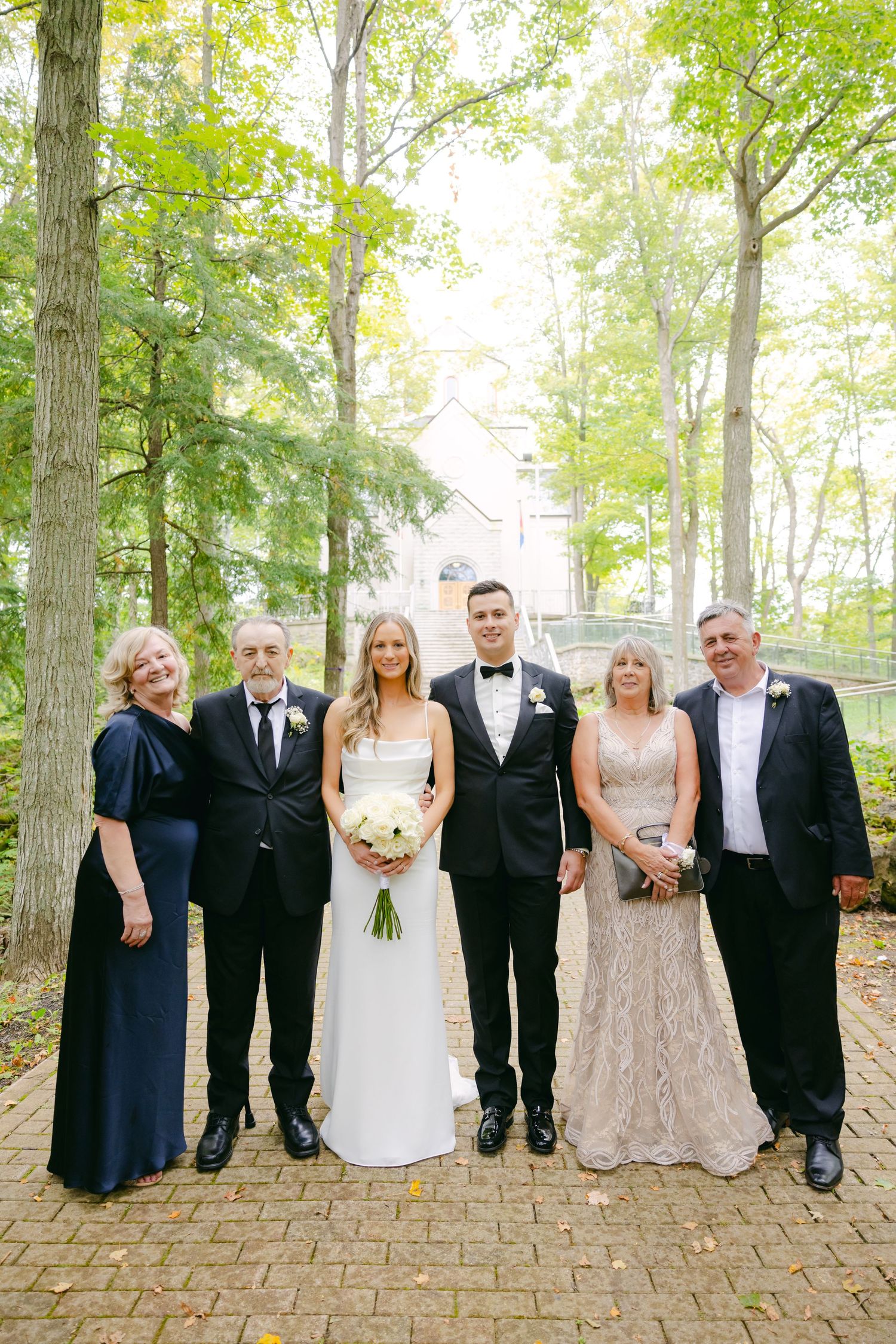 A wedding party poses for a formal family photo in a wooded outdoor setting.
