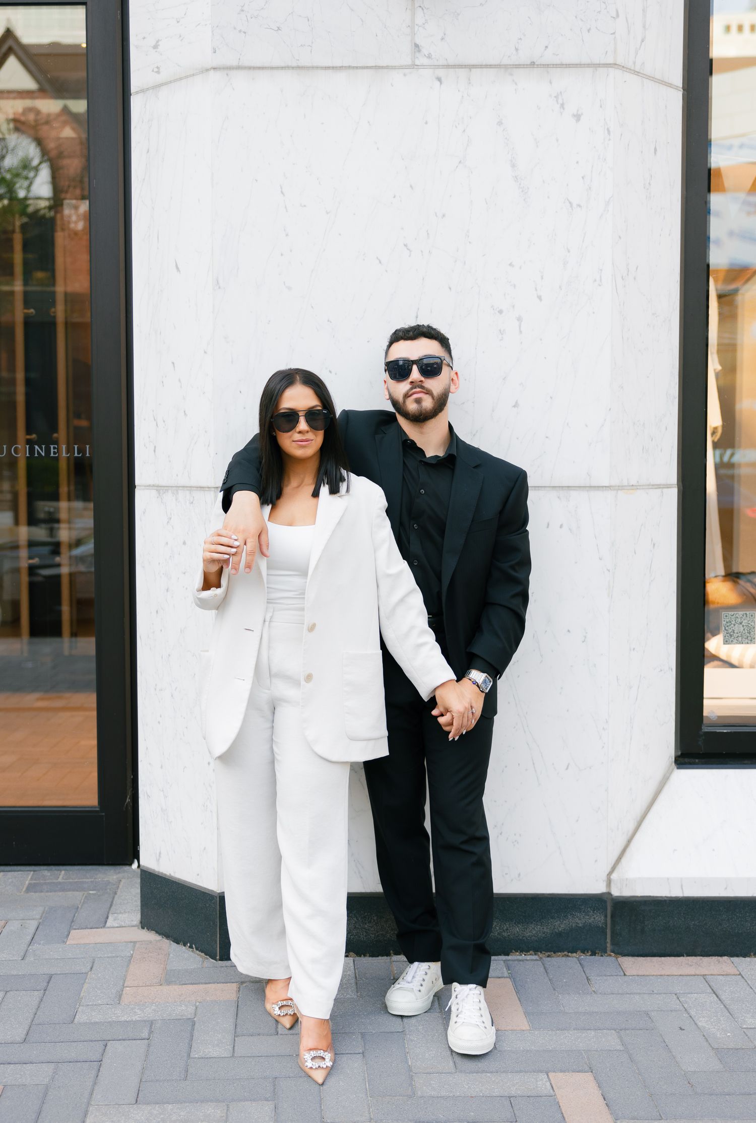 Modern couple in contrasting black and white suits pose together against white building exterior.