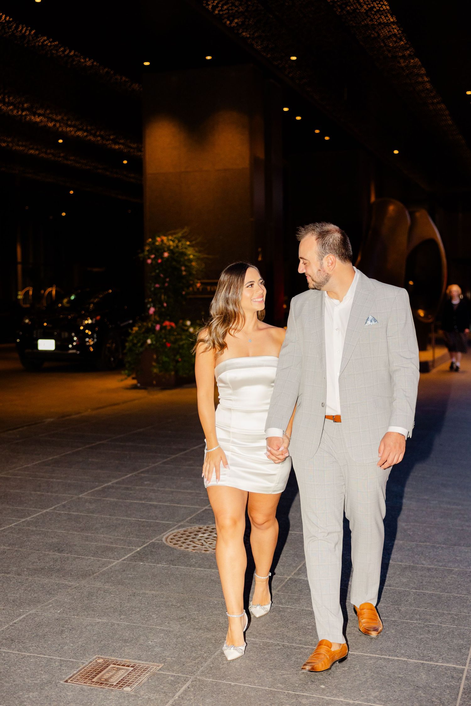 Couple walks together on city plaza wearing formal attire at night.