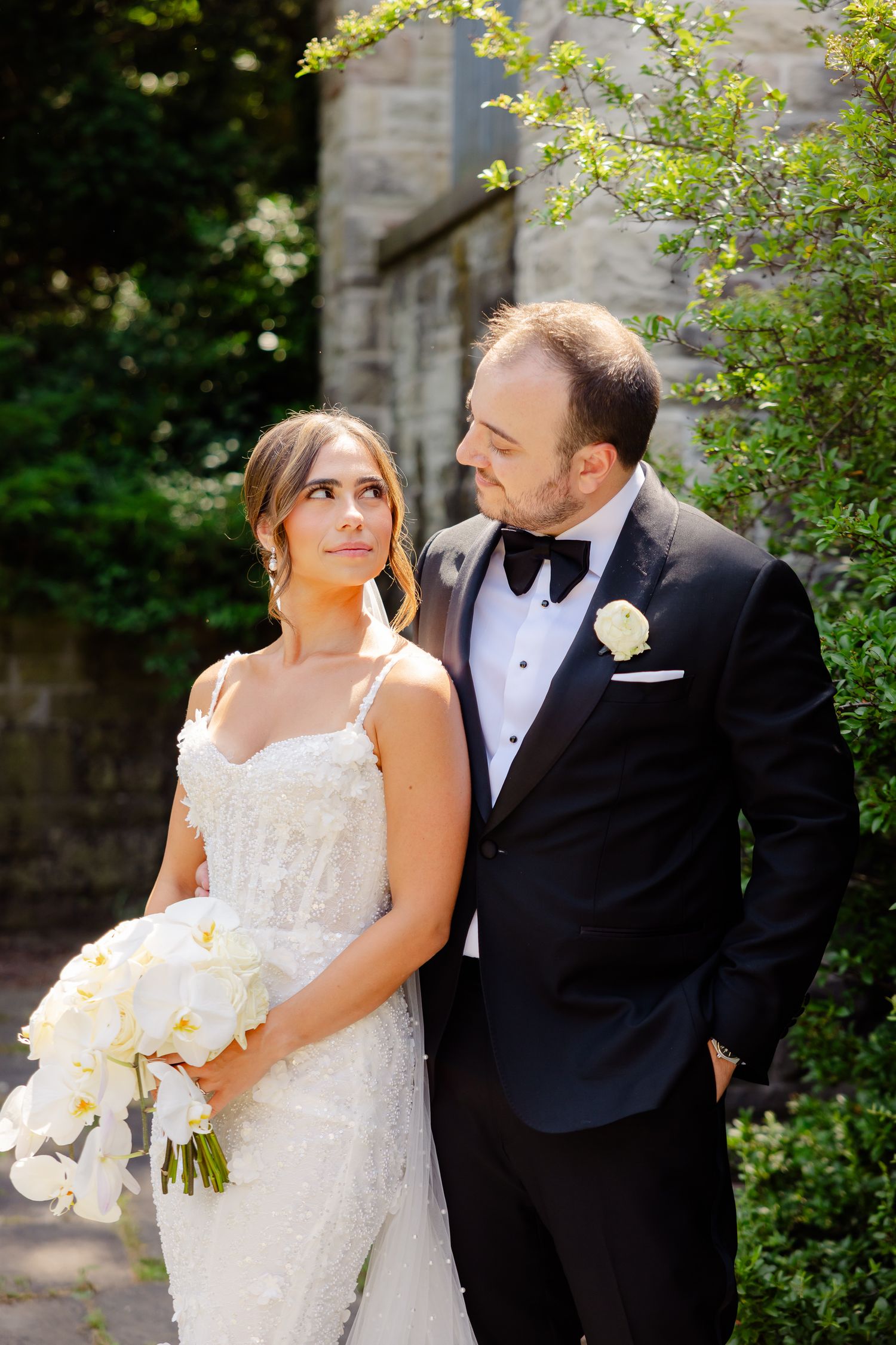 Elegant couple in wedding attire pose together outdoors by stone wall with greenery in formal garden setting.