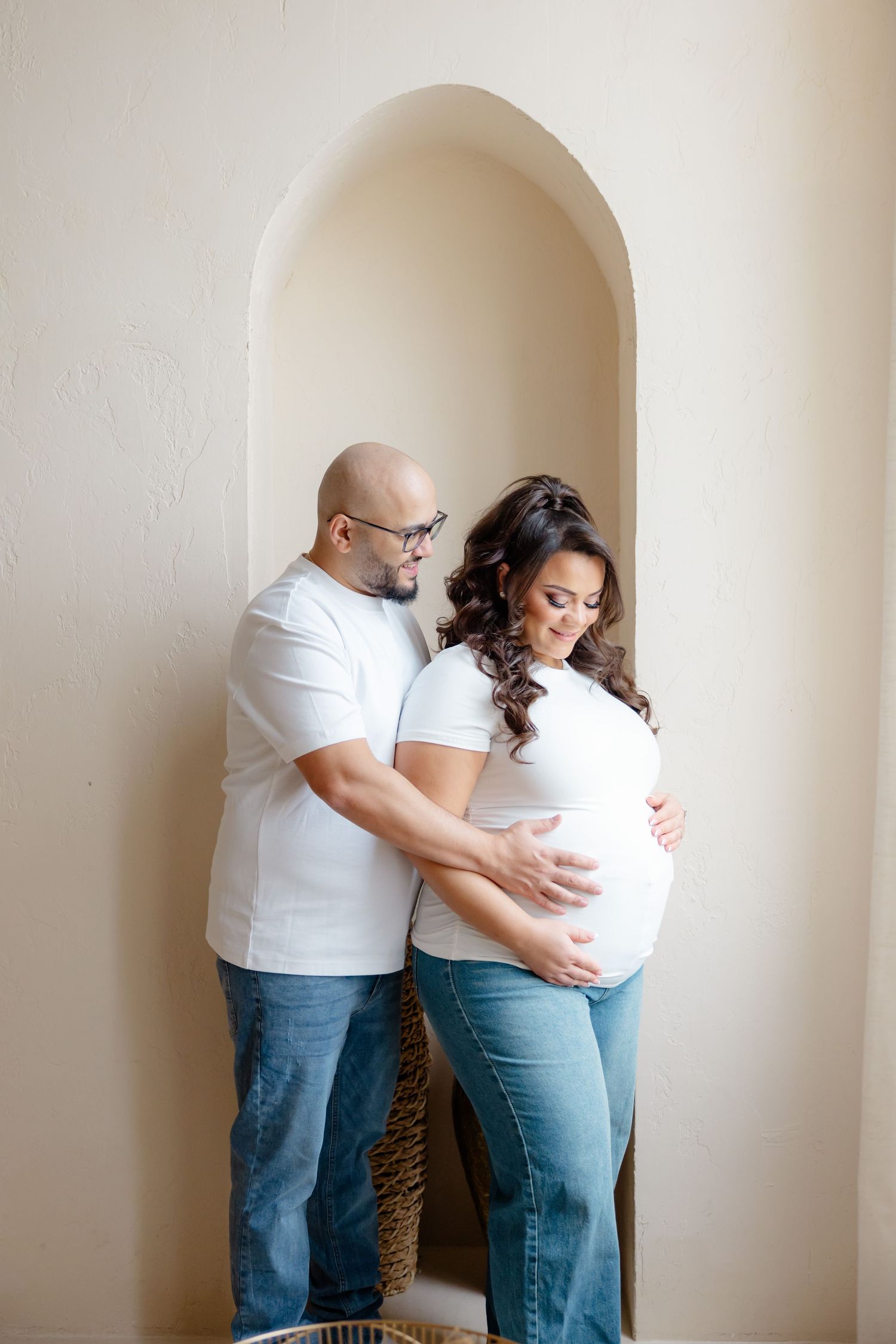 Expecting couple dressed in white shirts and jeans embrace while standing in an arched alcove.