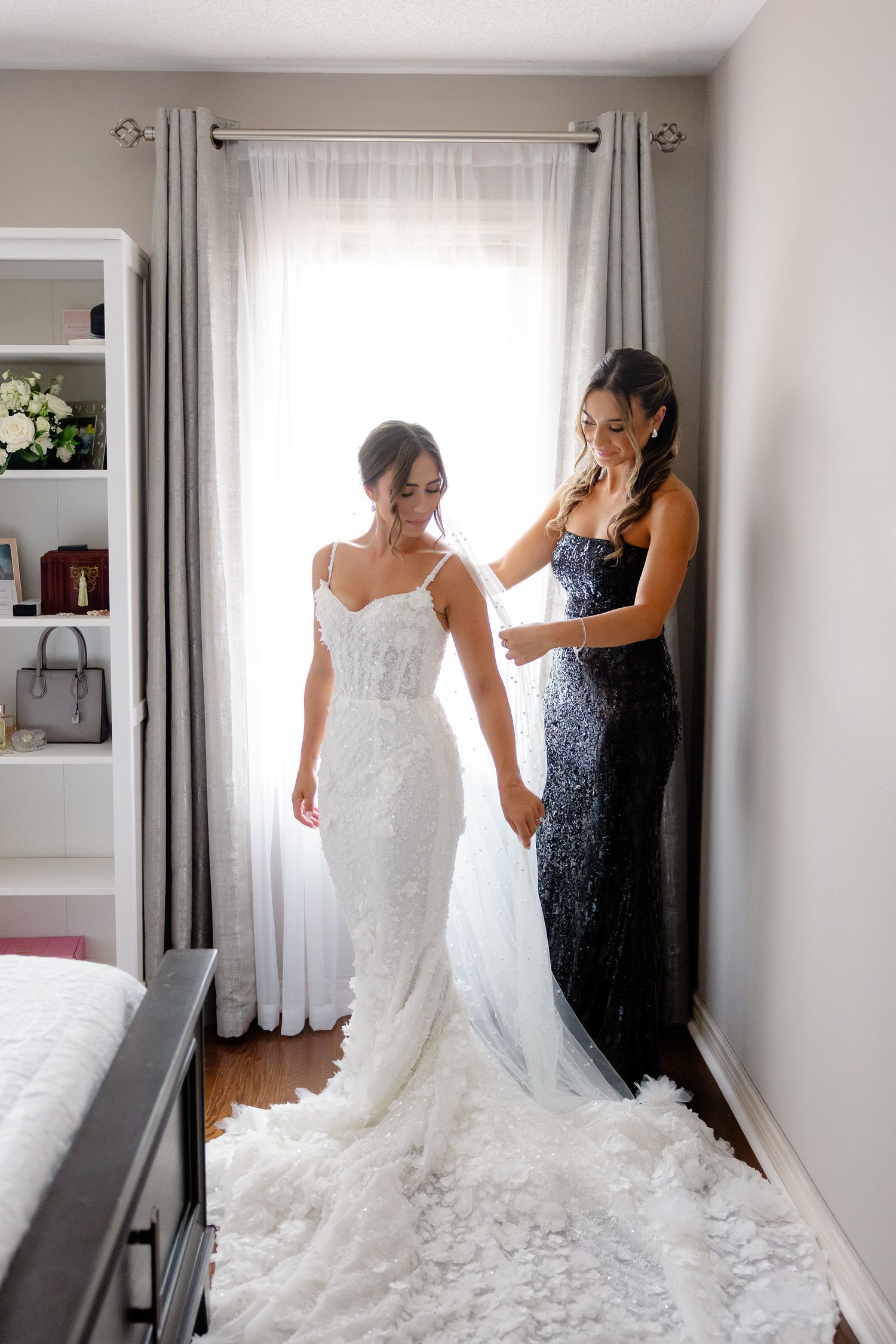 Someone in an elegant white wedding gown with a long train stands with another person wearing a navy sequined dress.