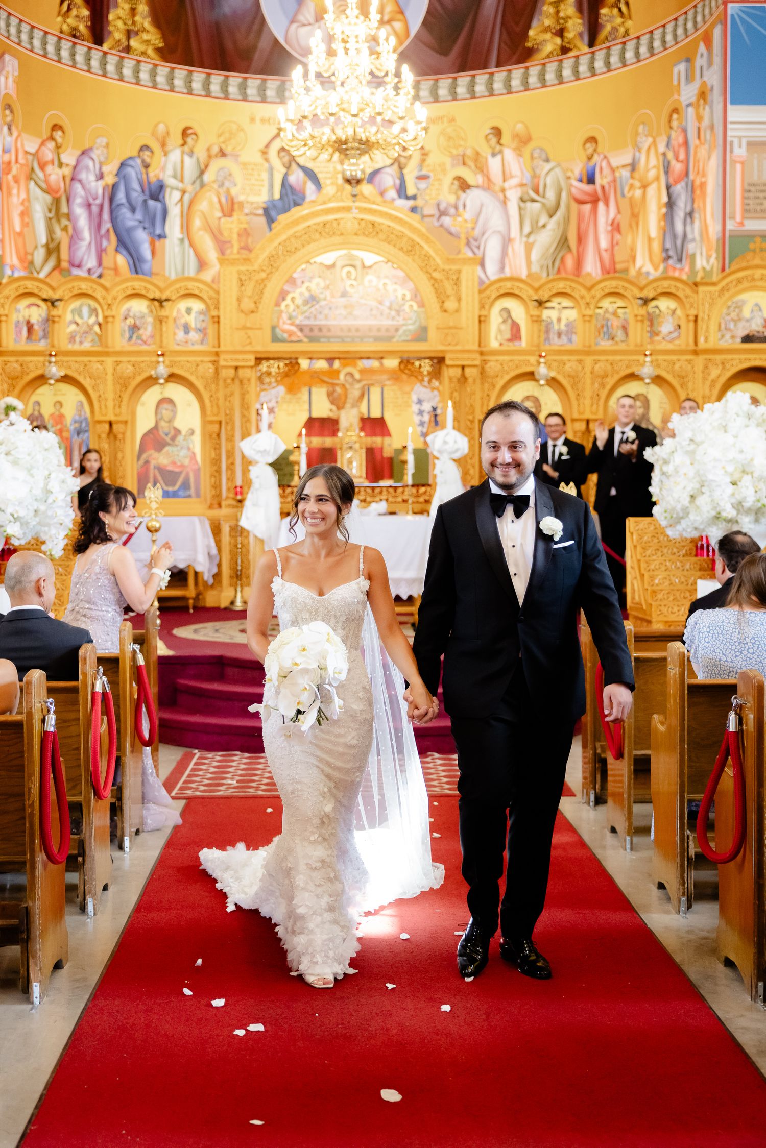 A wedding ceremony takes place in an ornate Orthodox church with golden religious murals and a red carpet aisle.