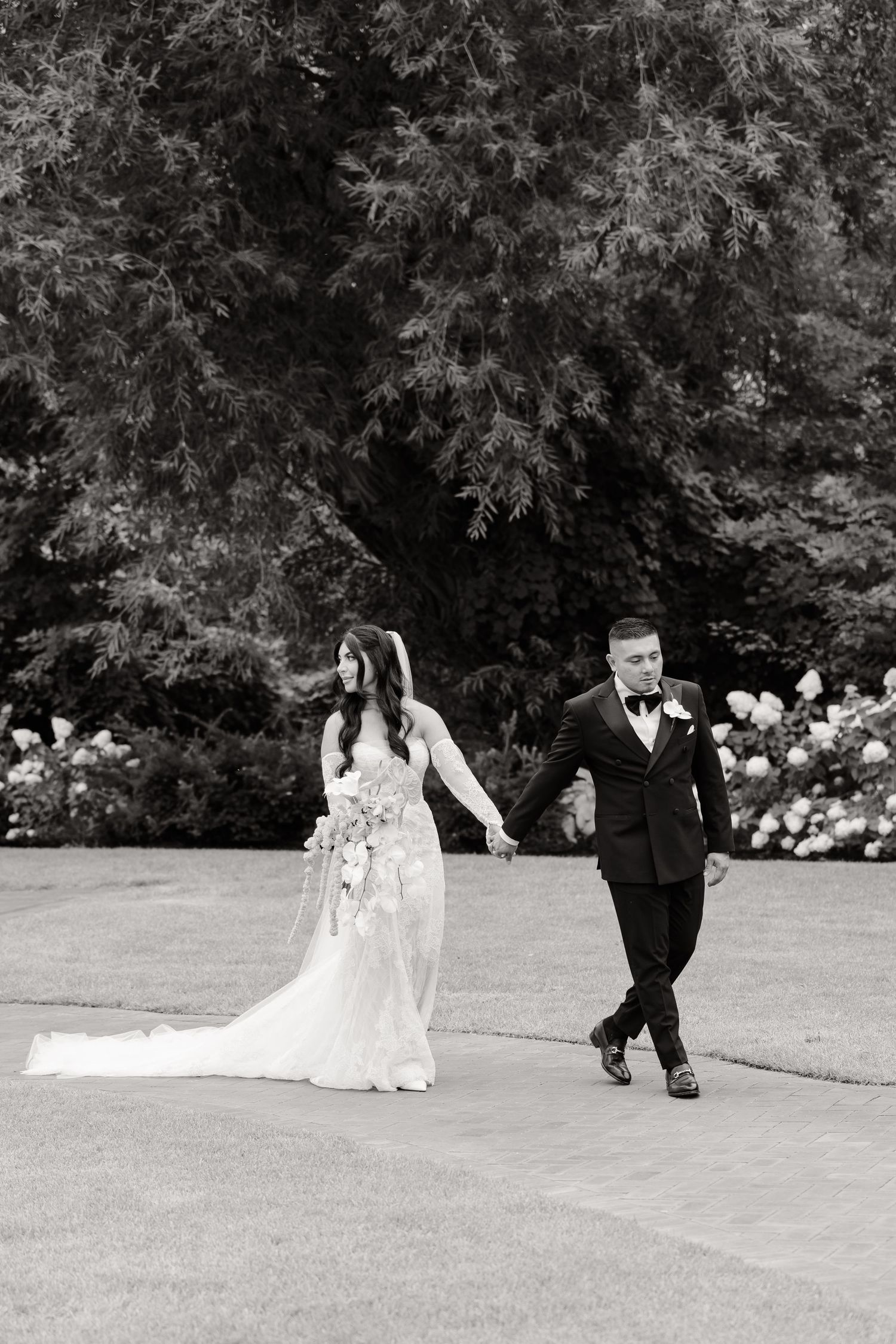 Wedding party members gracefully walk across a manicured lawn bordered by lush trees and blooming hydrangeas.