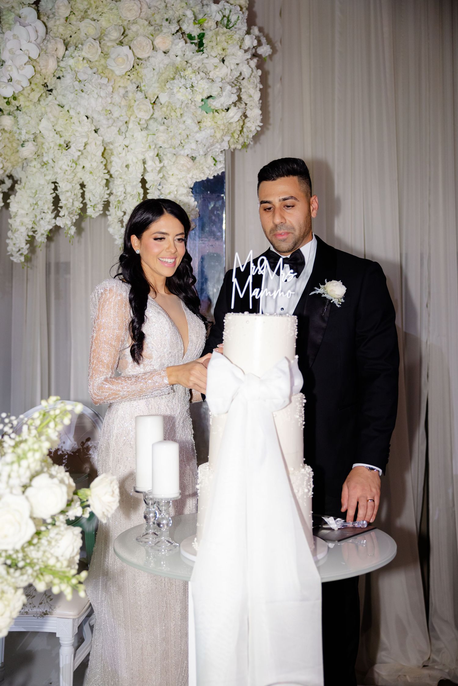 Couple poses with their modern white wedding cake amidst luxurious floral decorations.