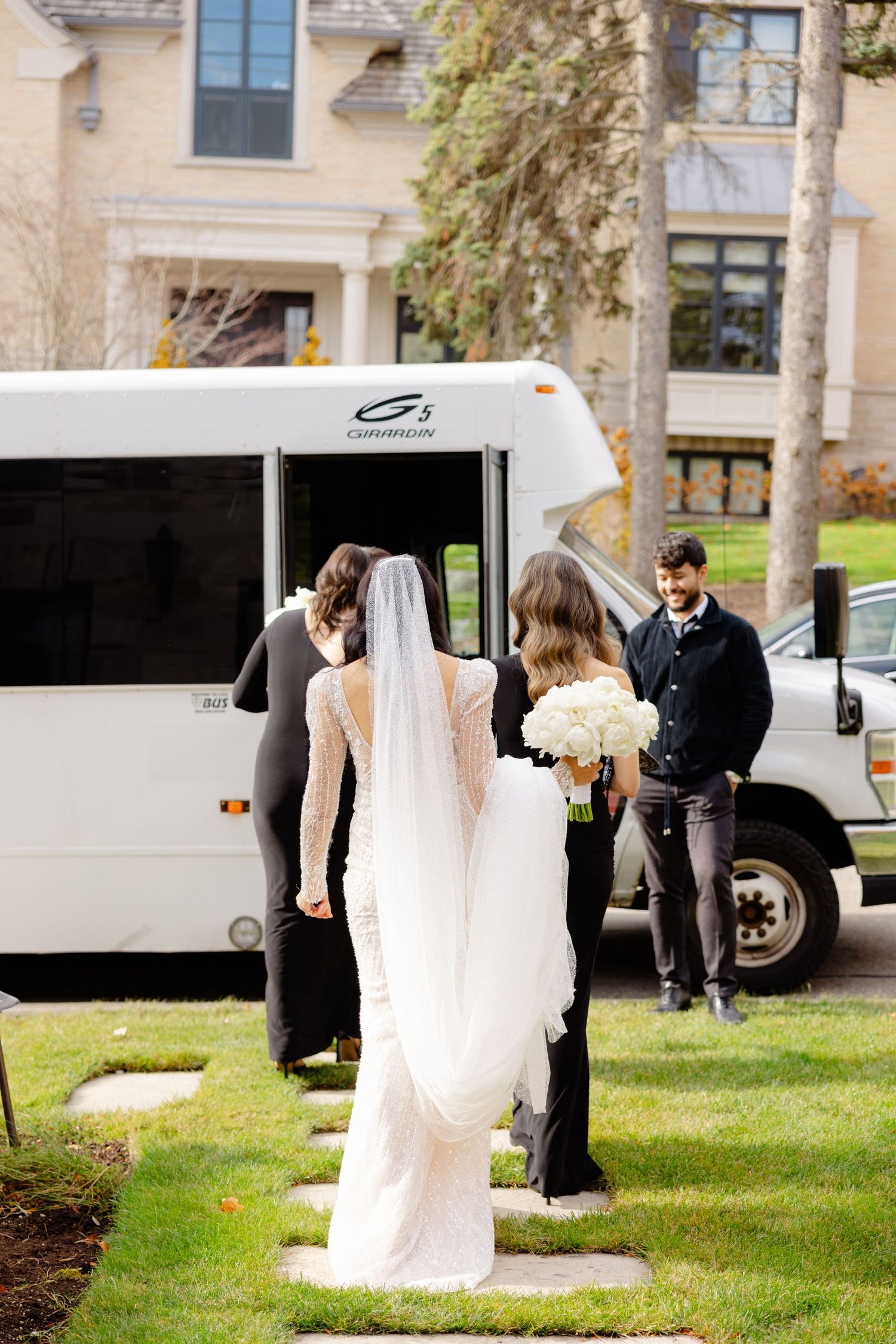 A bride in white walks towards a shuttle bus outside a building on her wedding day.