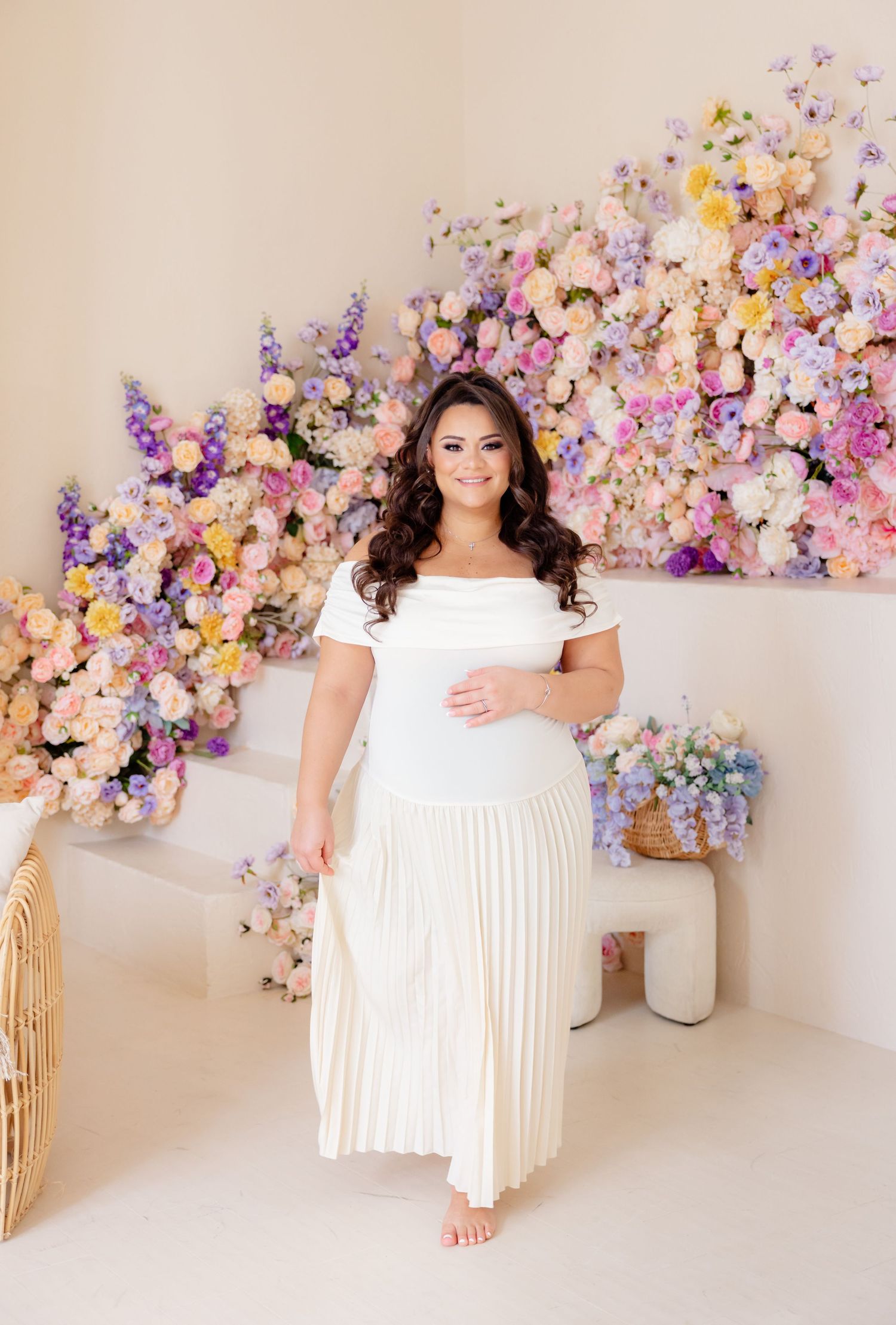 Elegant off-shoulder white pleated maxi dress photographed against a romantic floral backdrop of pink and purple blooms.