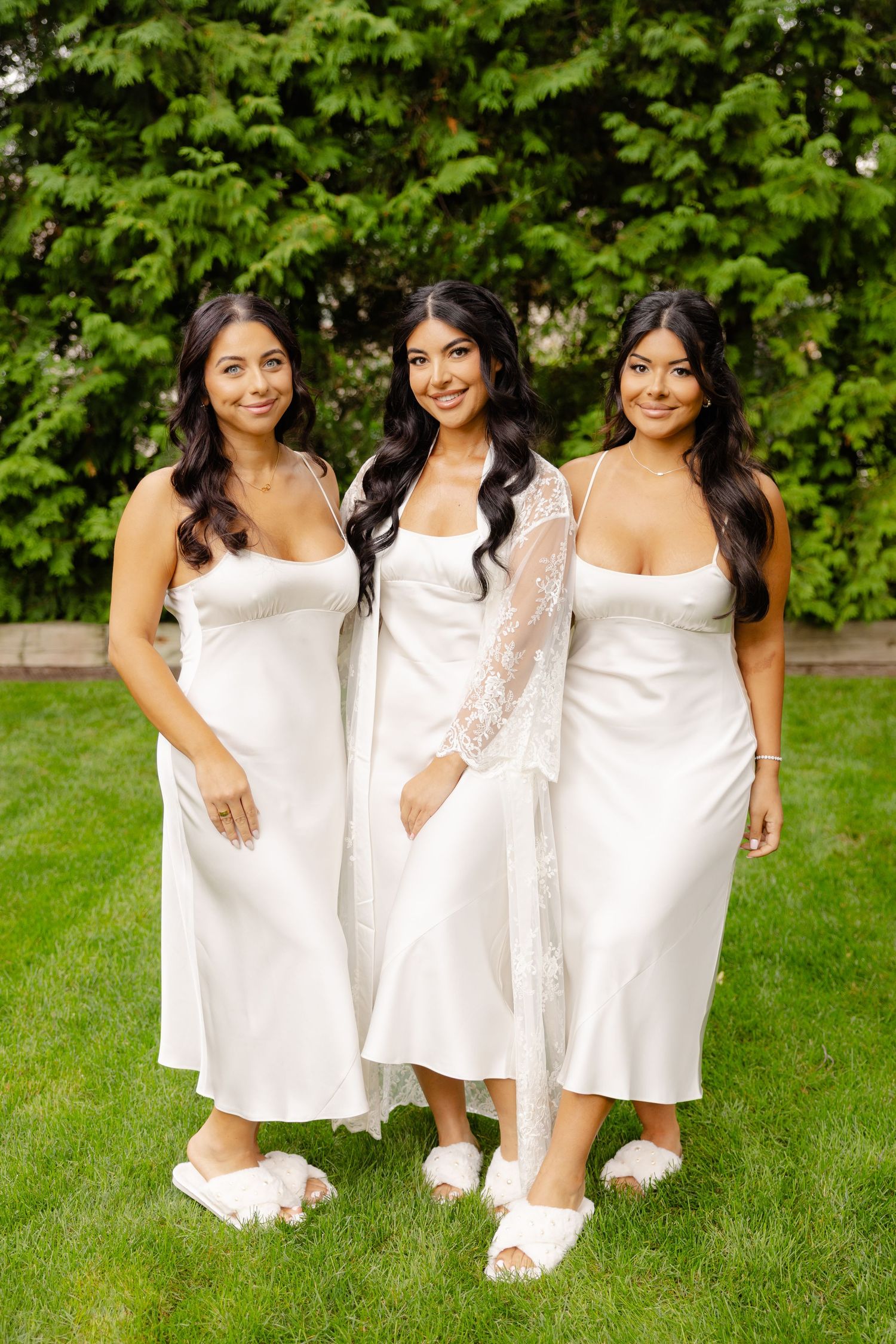 Three women in white dresses pose together on green grass with leafy foliage in the background.