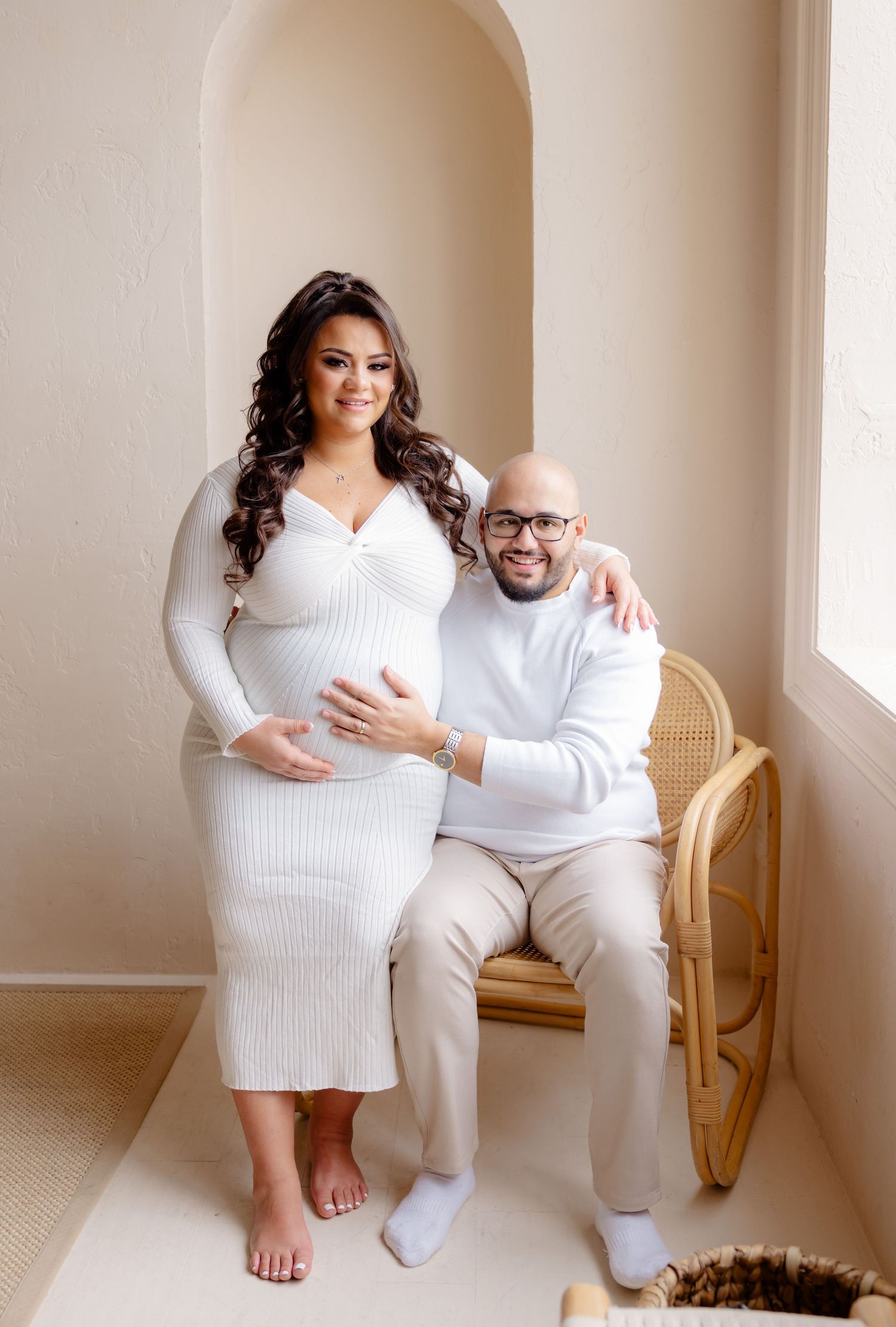 A couple in matching white outfits pose together in an arched alcove with a rattan chair during a maternity photoshoot.
