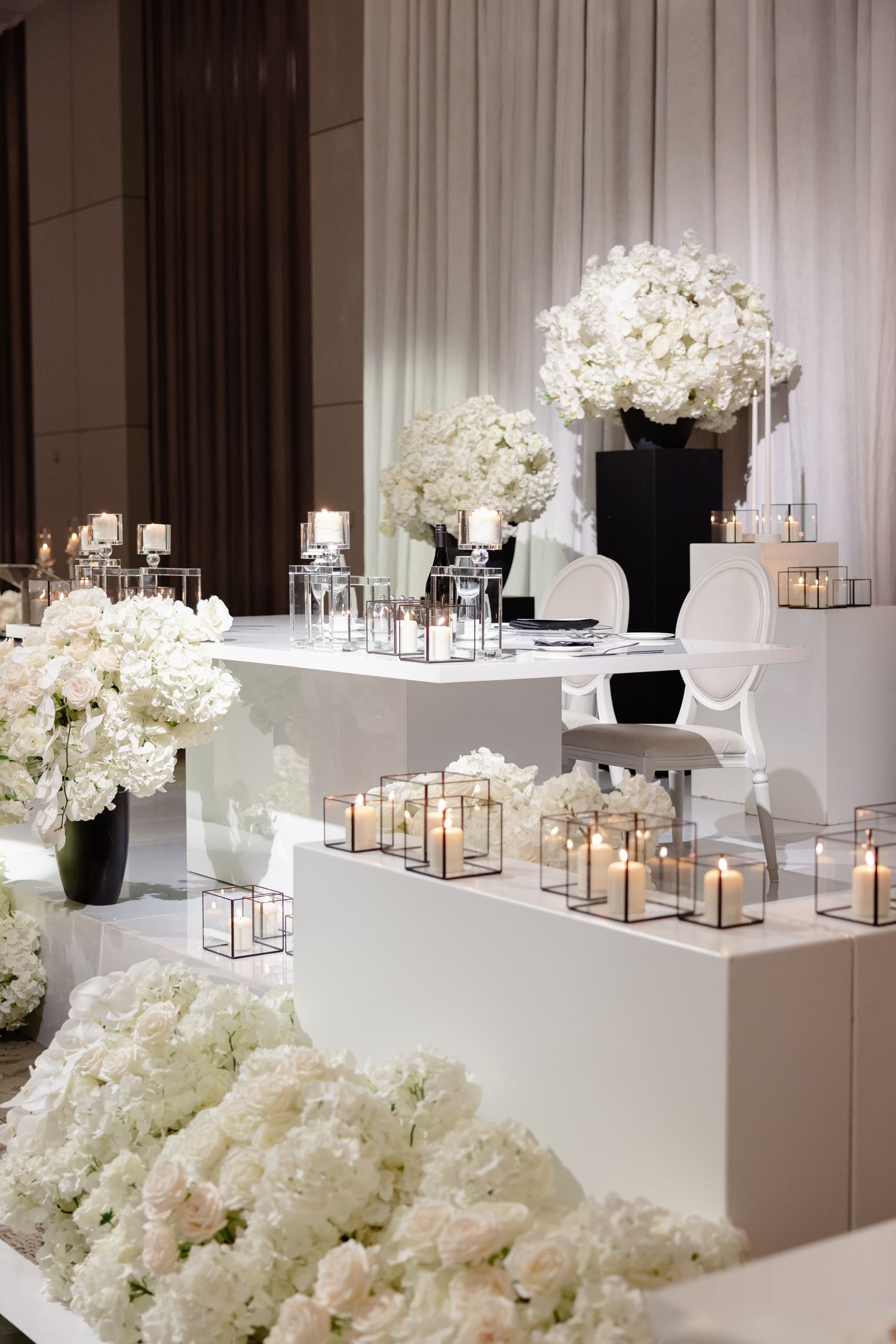 Elegant wedding reception setup with white hydrangeas and romantic candle lanterns on tiered platforms.