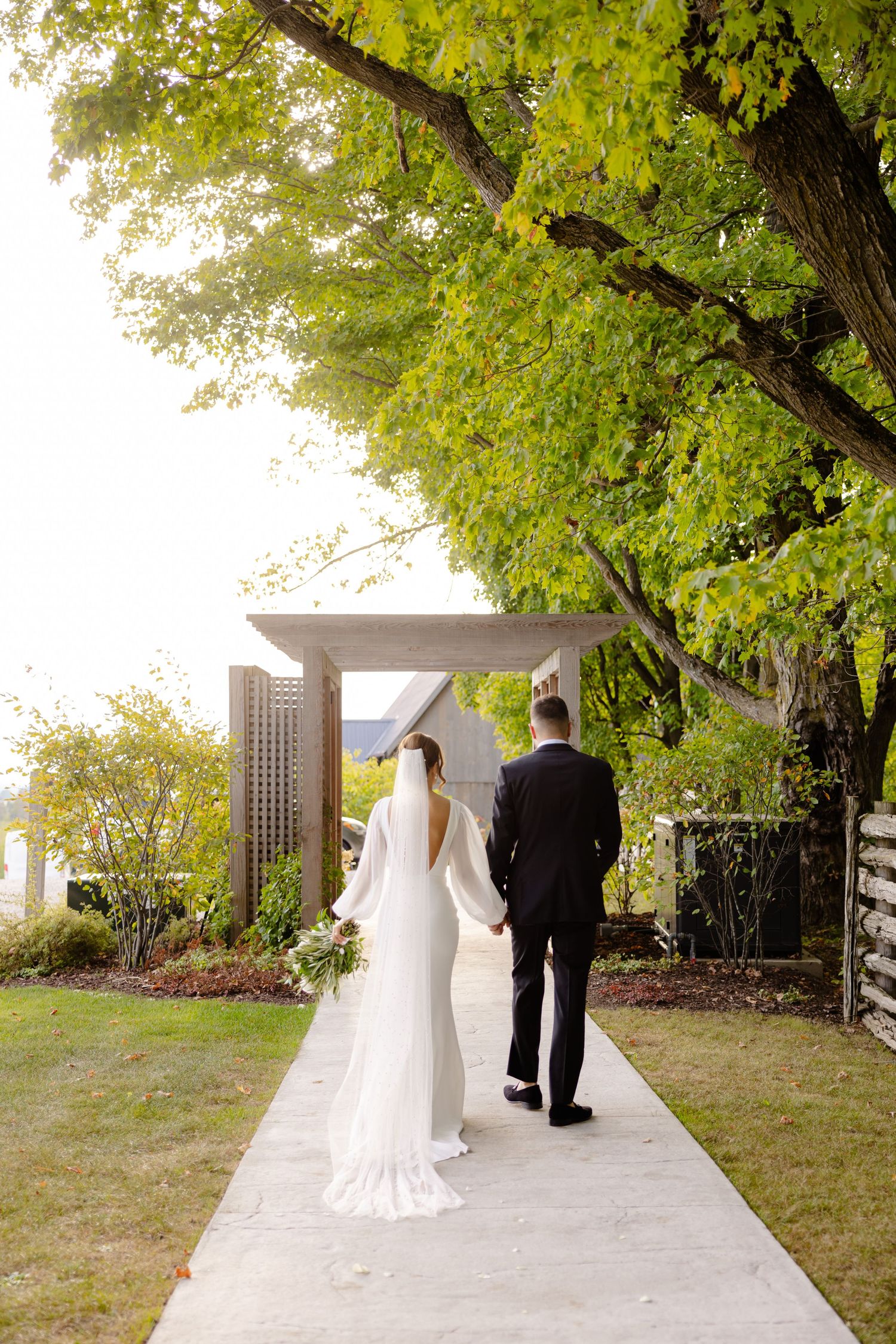 Newlyweds walking down a garden path under lush green trees on their wedding day.