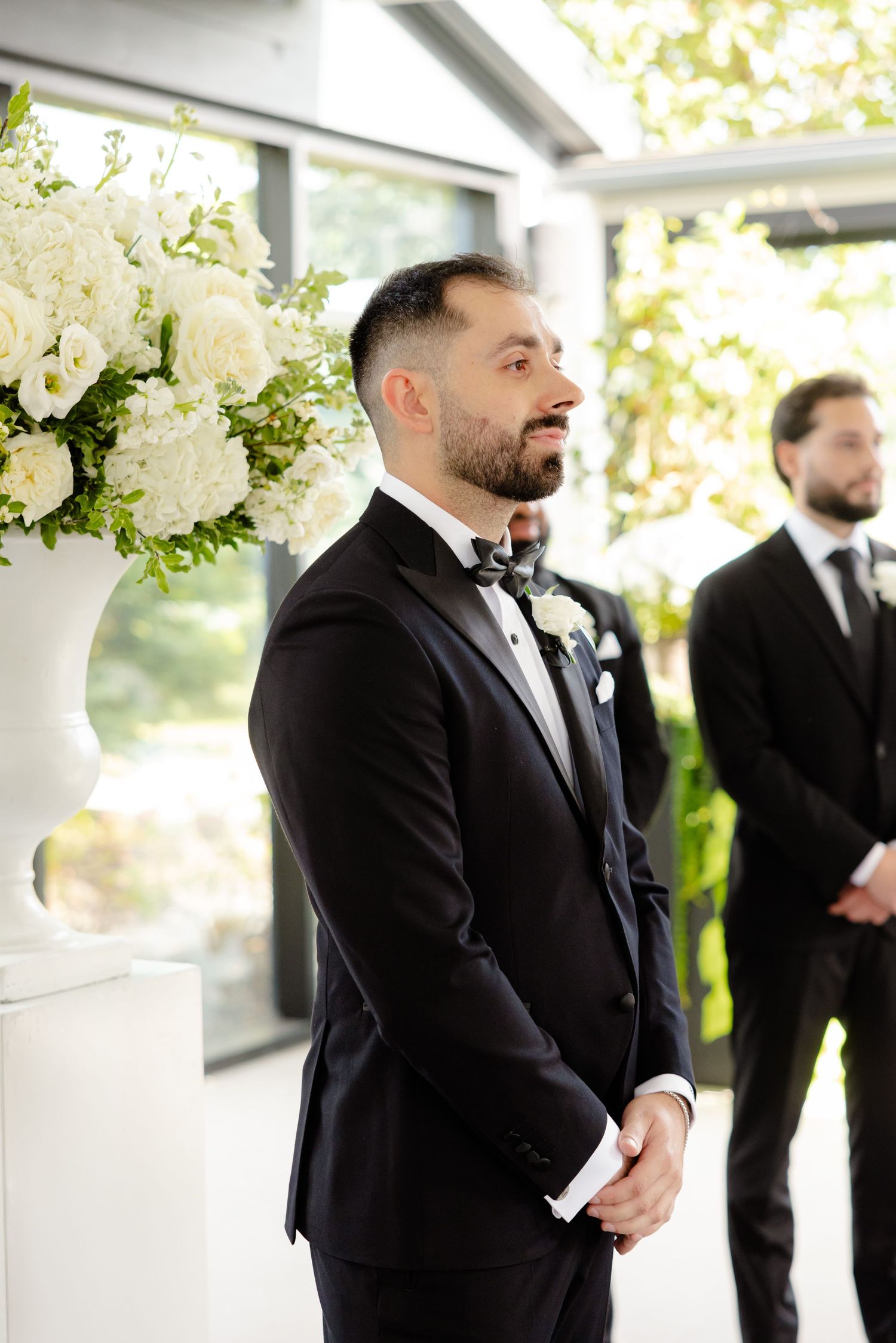 A groom stands at the altar beside white floral arrangements in a sun-filled wedding venue.