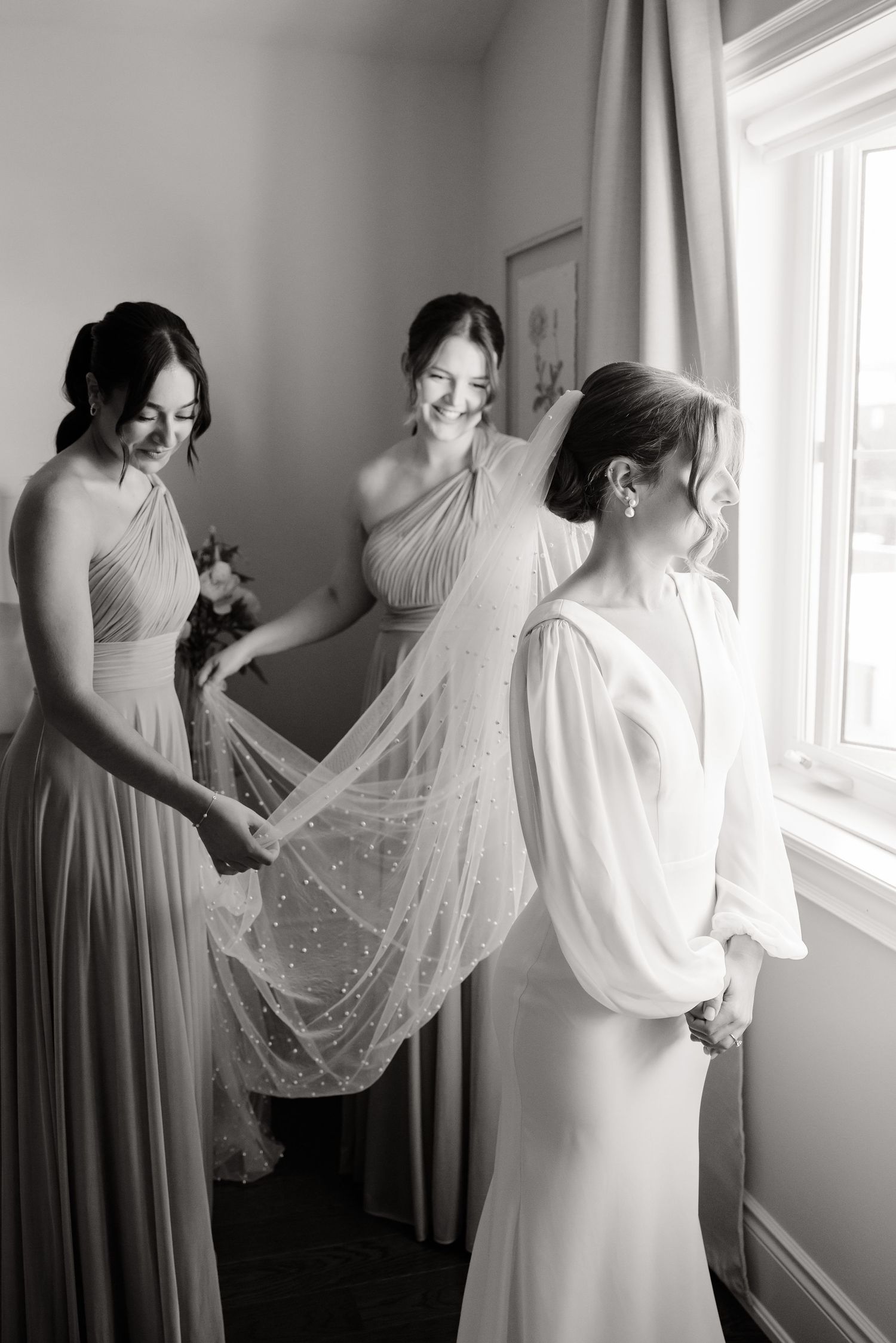 Bridesmaids in sage dresses help with bridal veil during wedding day preparations.