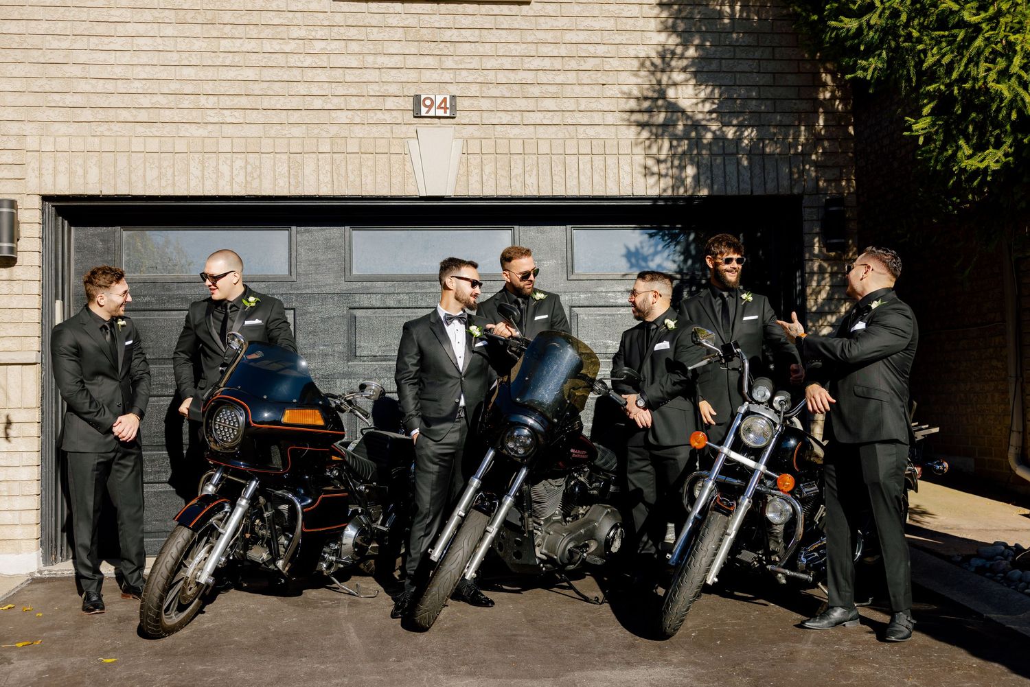 Several motorcycles and riders gathered in front of a beige building's garage.