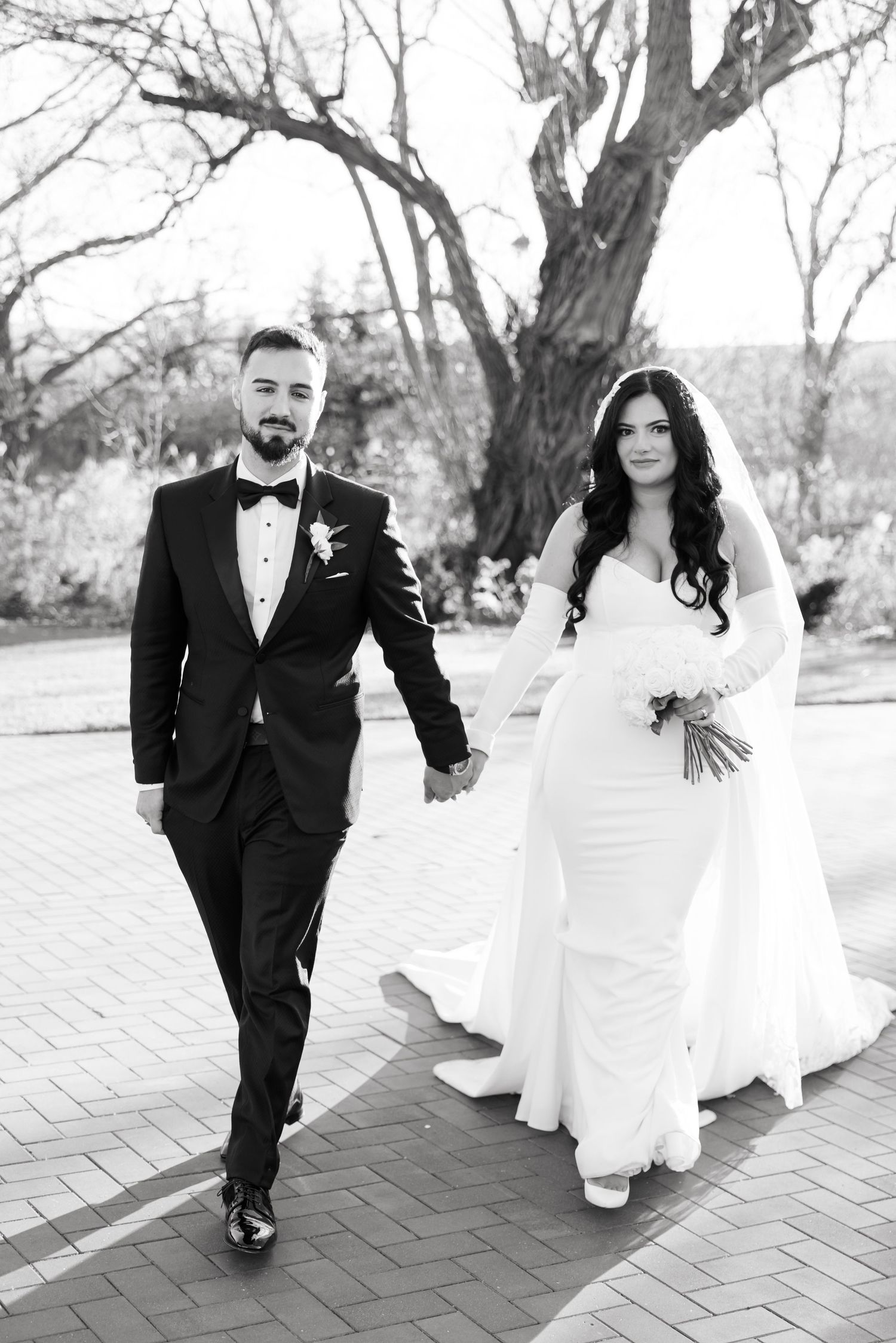 A black and white wedding photo showing a couple walking together on a brick pathway beneath a bare tree.