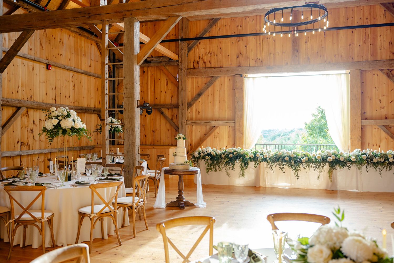 A rustic barn wedding reception setup with wooden crossback chairs and white floral decorations.