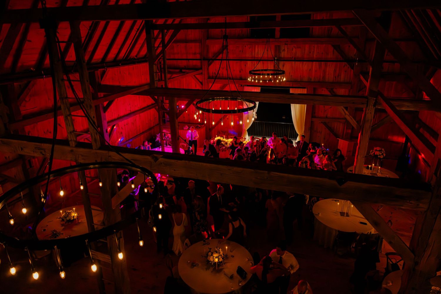 Dramatic red-lit rustic barn wedding venue with hanging string lights and chandelier.
