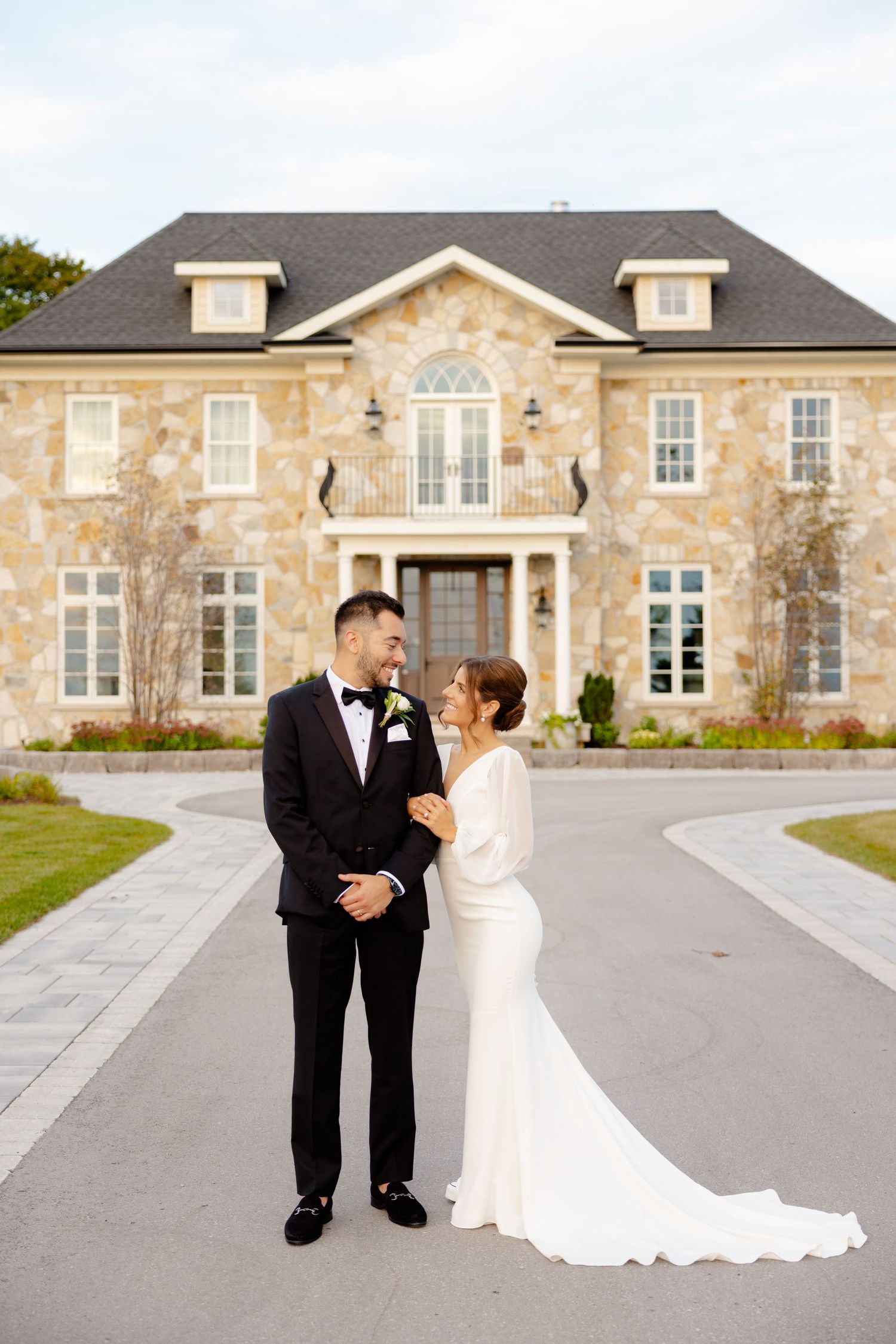 A couple poses in wedding attire in front of a luxurious stone mansion.