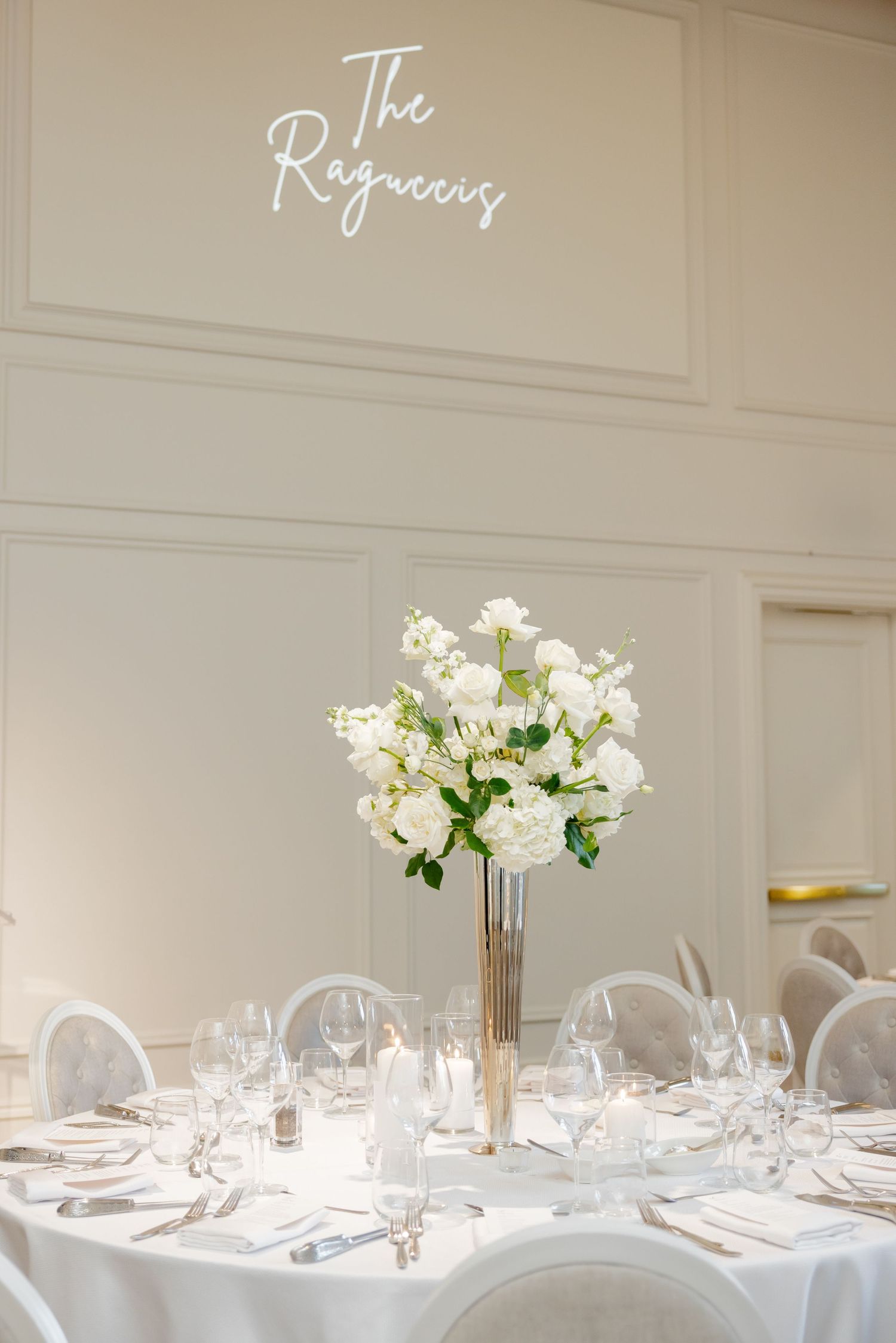 White wedding reception table under 'The Regency' sign with tall floral centerpiece and place settings.
