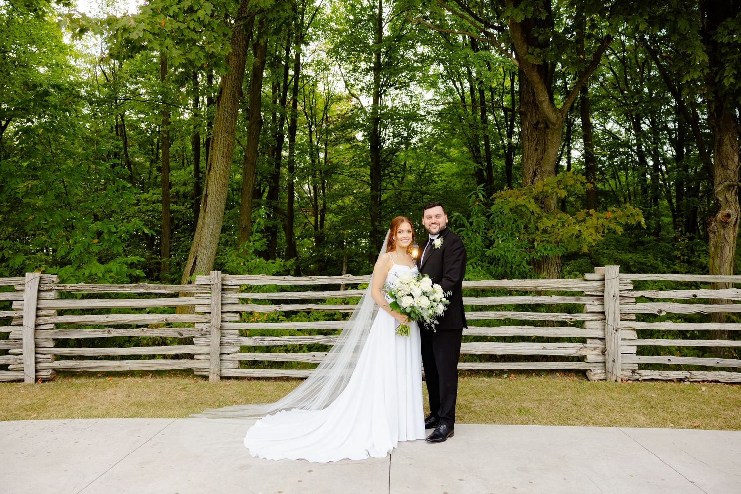 Wedding portrait in front of rustic wooden fence with forest backdrop.