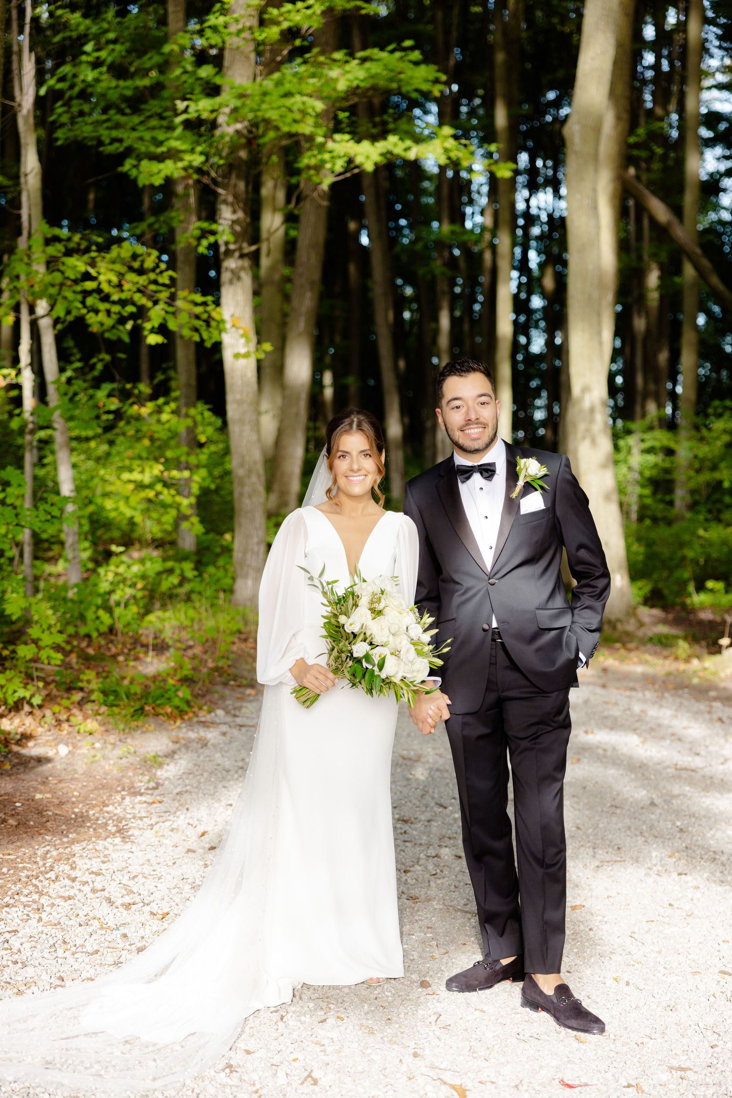 Bride in elegant white dress and groom in tuxedo pose on forest path during summer wedding.