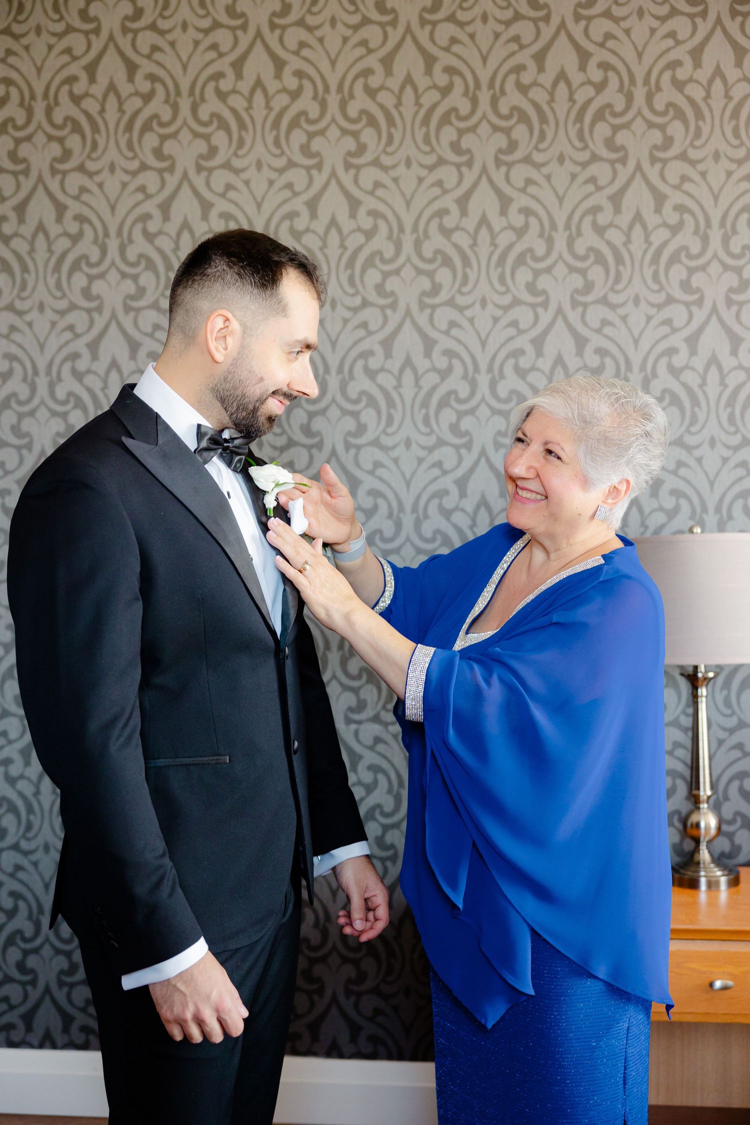 A woman in blue helps adjust a boutonniere on a person's tuxedo lapel against a patterned wall.