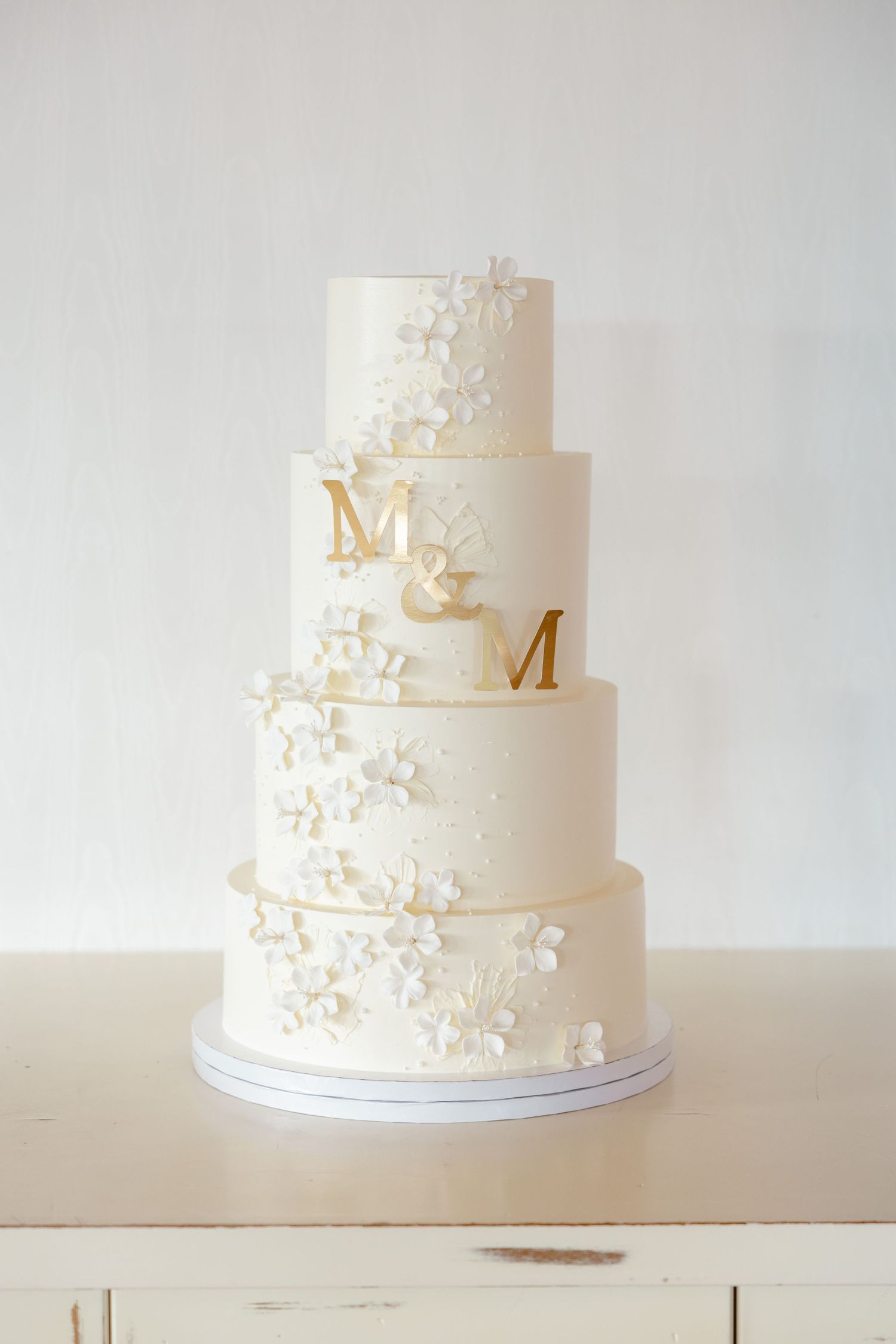 Four-tier white wedding cake decorated with delicate floral details and gold M letters.