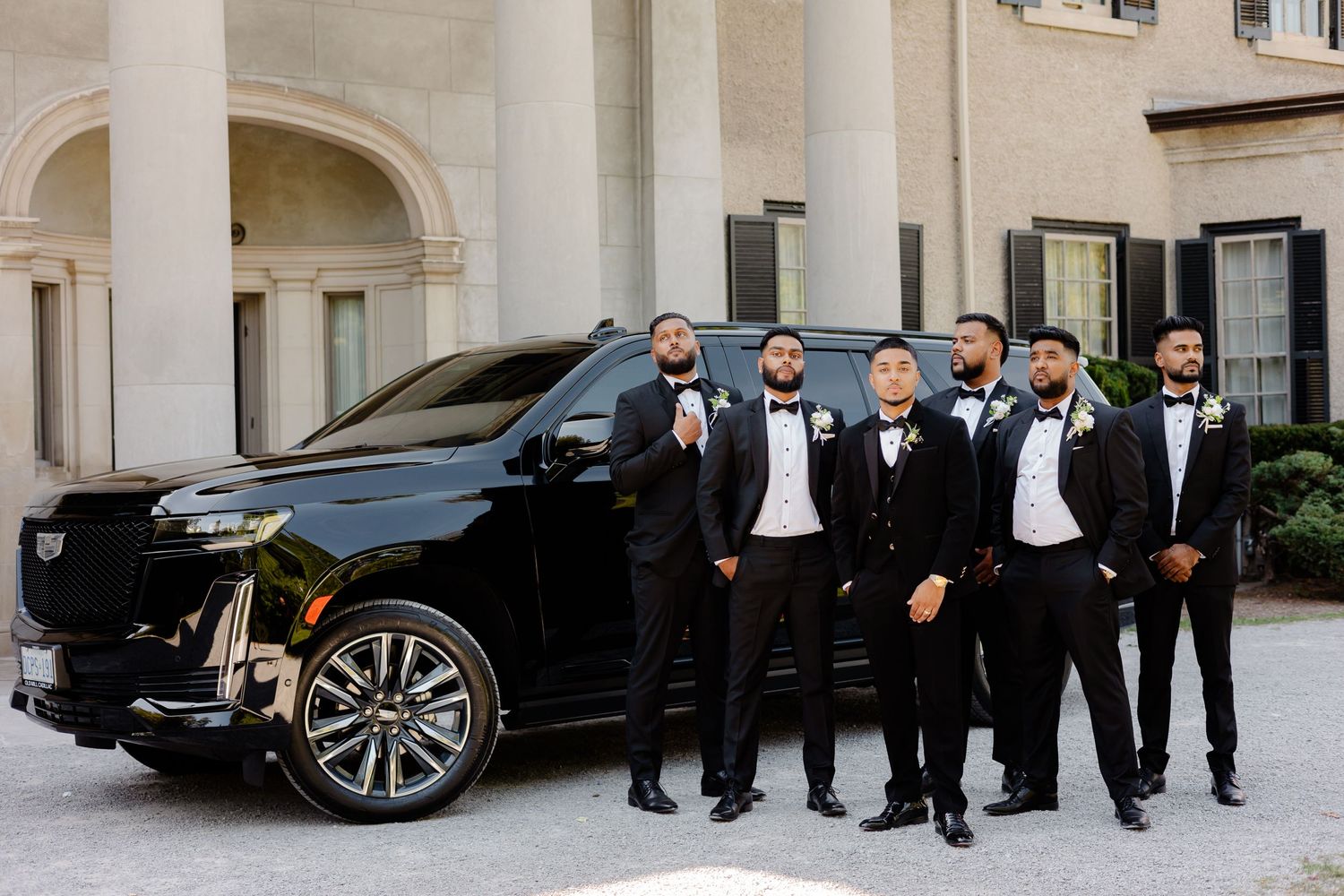 Wedding party of groomsmen in tuxedos posing with a black luxury SUV.